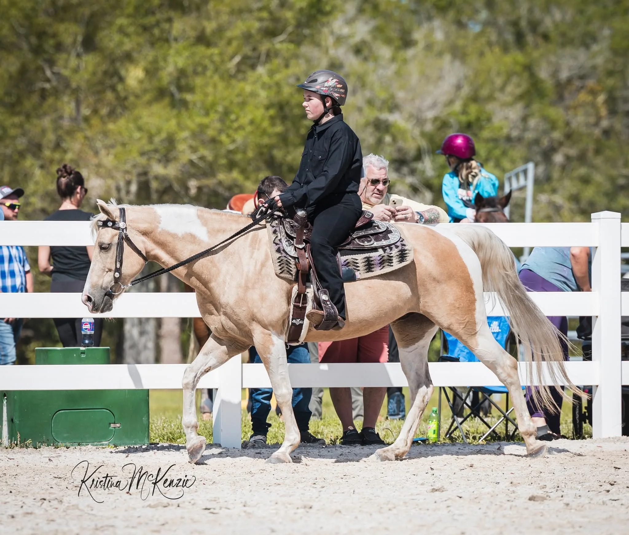 A young woman riding a horse during a competition or riding event, with people in the background and lush green trees.