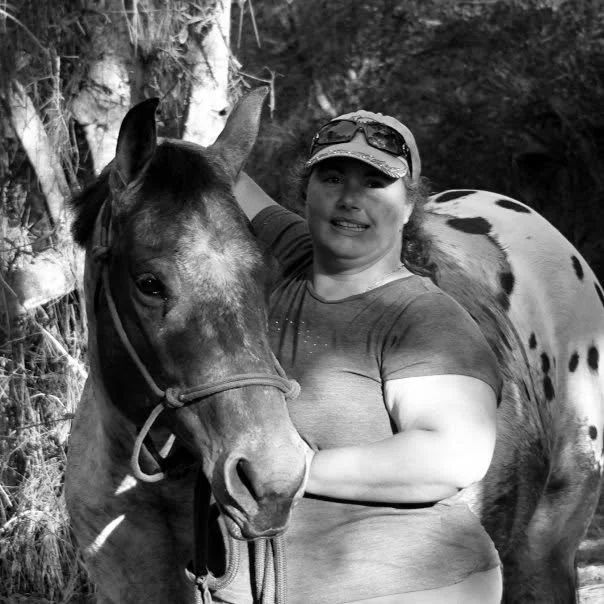 A woman with sunglasses and a cap standing beside a horse with a spotted coat in an outdoor wooded area.