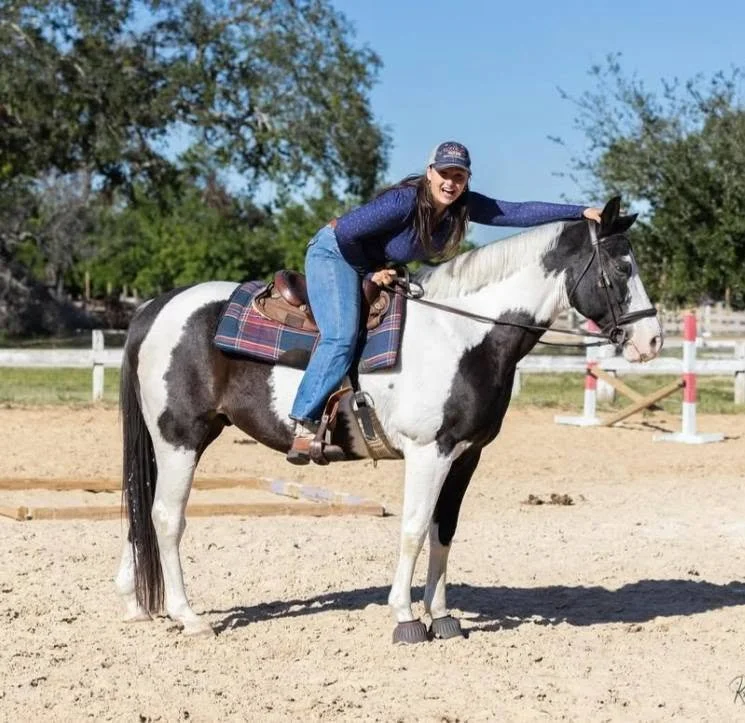 Woman riding a black and white horse on a dirt riding arena, with green trees and a clear blue sky in the background.