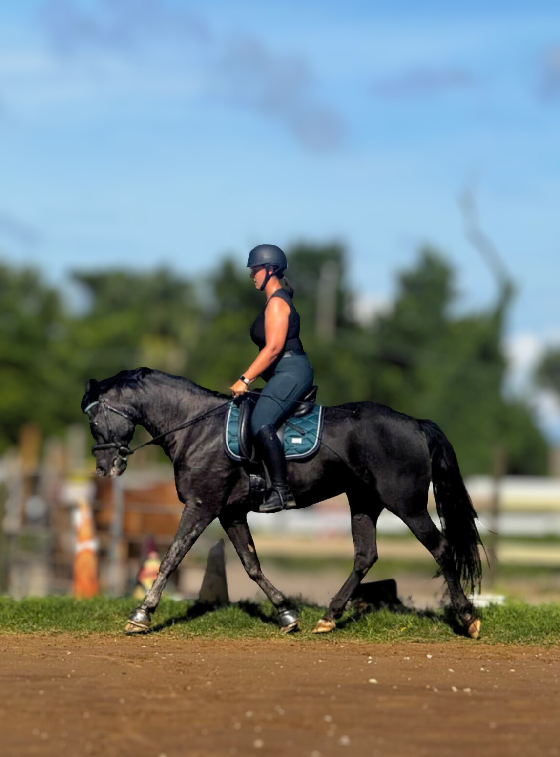A woman riding a black horse in an outdoor equestrian arena, wearing a helmet, black sleeveless top, riding pants, and tall riding boots, with trees and a blue sky in the background.