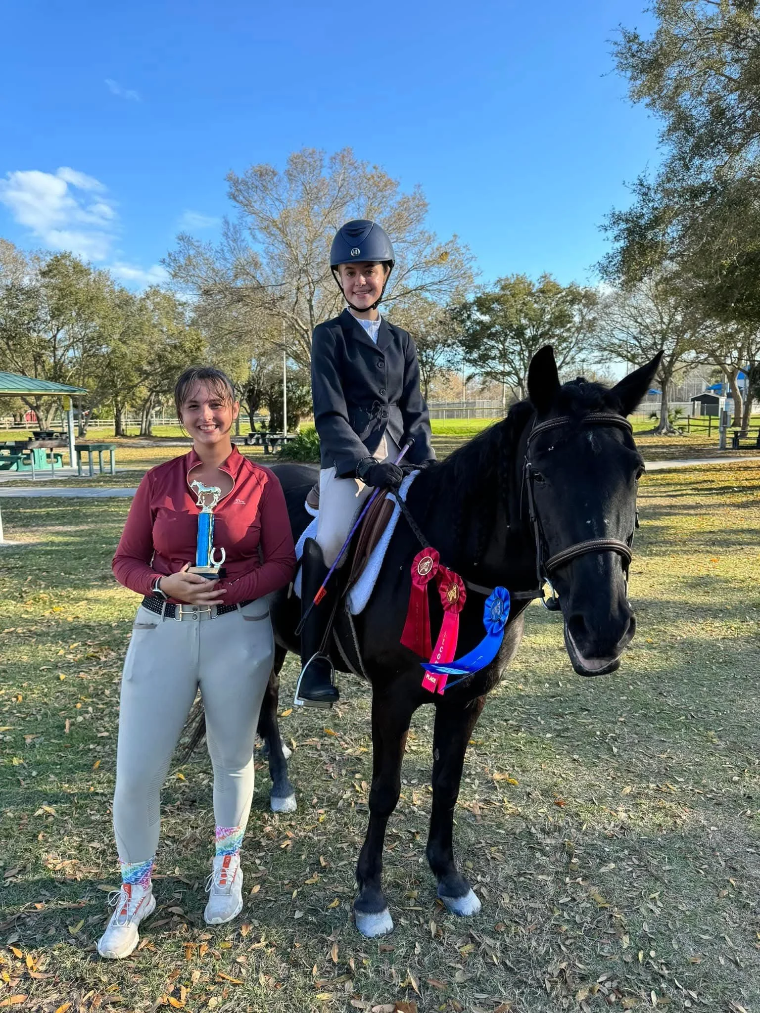 Two young girls and a black horse in a park with trees and a pavilion in the background. One girl is standing and holding a trophy, smiling, dressed in a reddish jacket and white riding pants. The other girl is sitting on the horse, wearing a helmet,