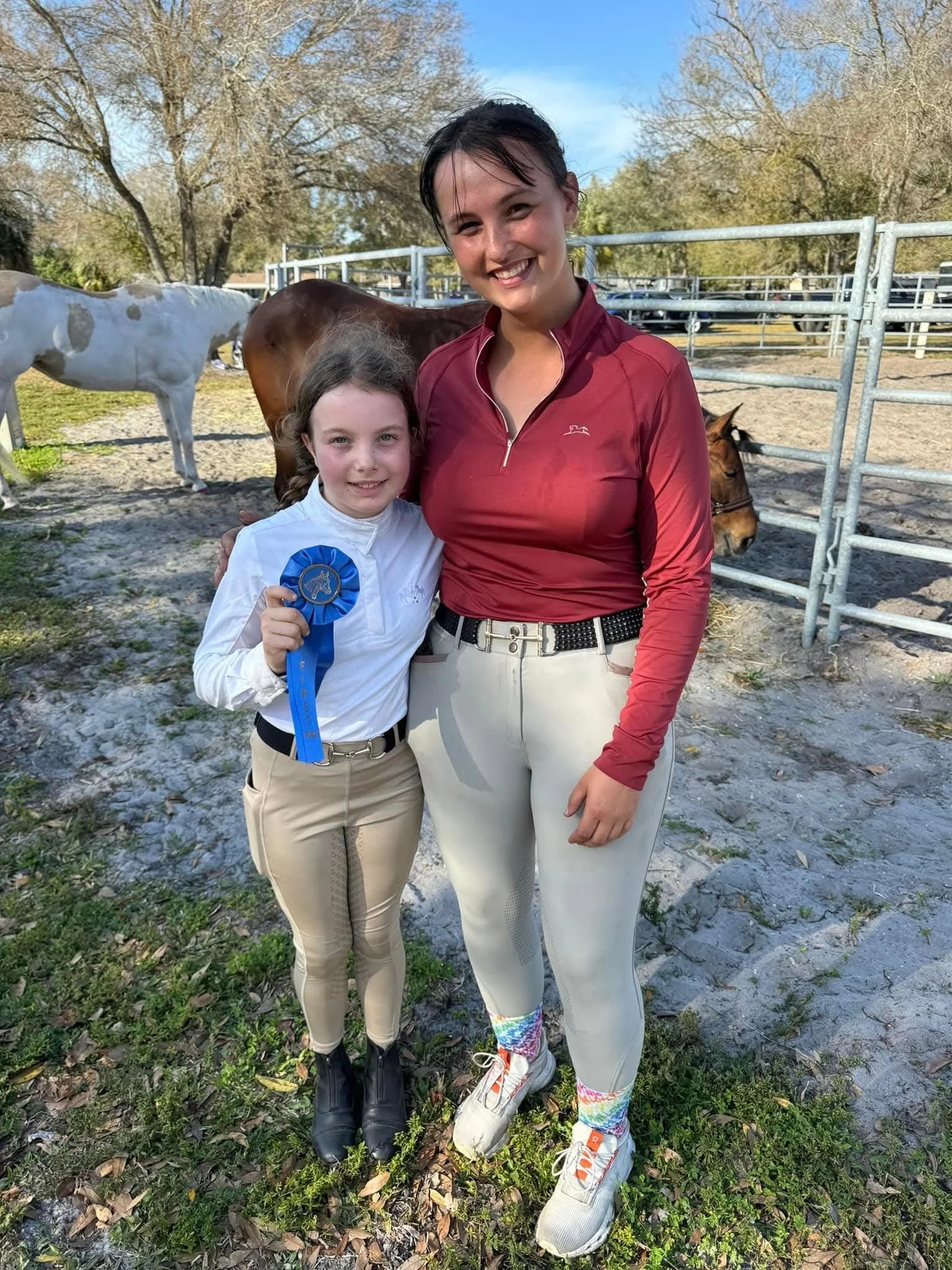 Two smiling girls posing together outdoors with horses and trees in the background. One girl is holding a blue ribbon. The photo is taken on a clear day with green grass and leaves on the ground.