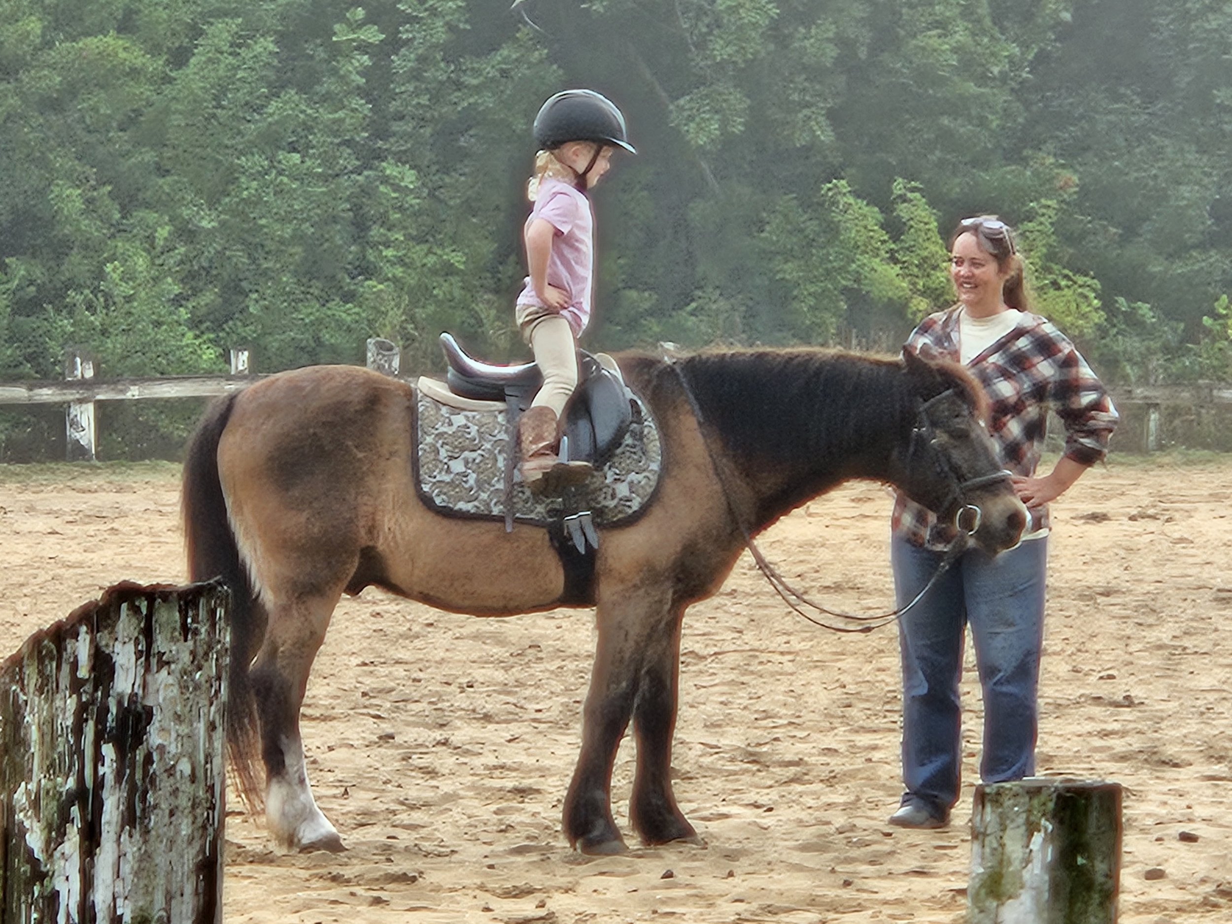 A young girl riding a pony horse with an adult woman standing nearby, smiling, outdoors in a fenced area with trees in the background.