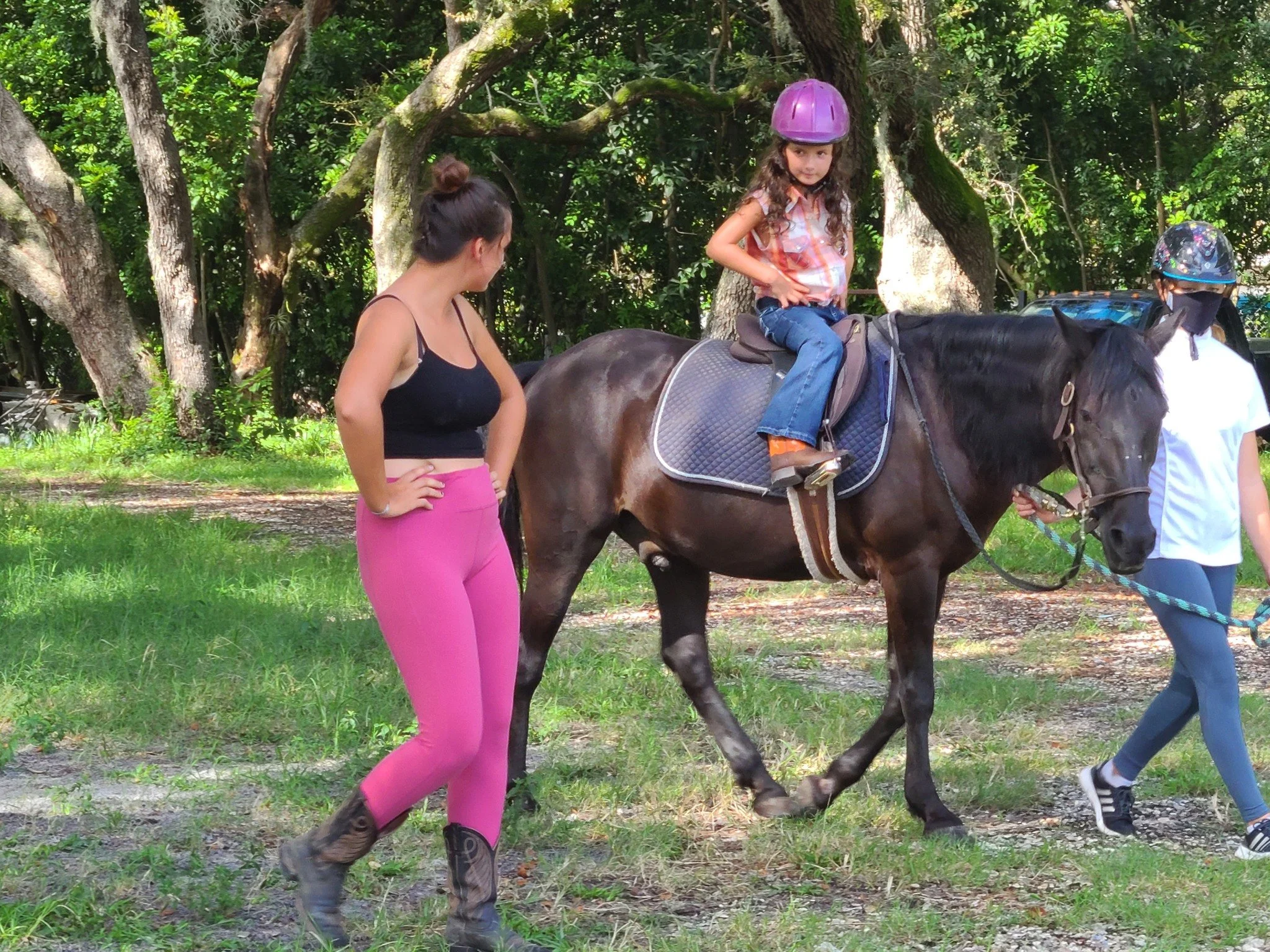 A young girl with curly hair sitting on a brown horse, wearing a purple helmet and a pink and orange plaid shirt. An adult woman with dark hair tied back, wearing a black tank top and pink pants, stands nearby. Another person with a helmet and face m