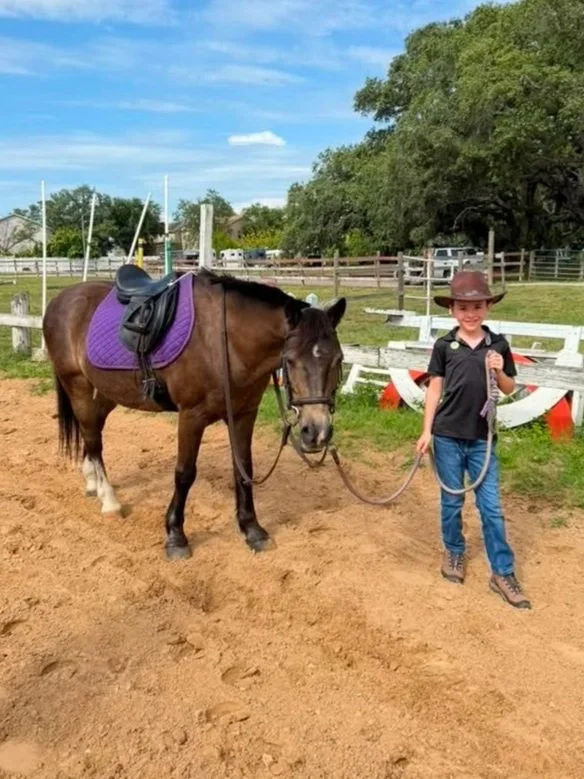 A young boy wearing a cowboy hat, black shirt, and jeans holding a lead rope attached to a brown horse with a purple saddle pad and black saddle in an outdoor riding arena with fencing, trees, and a blue sky with clouds.