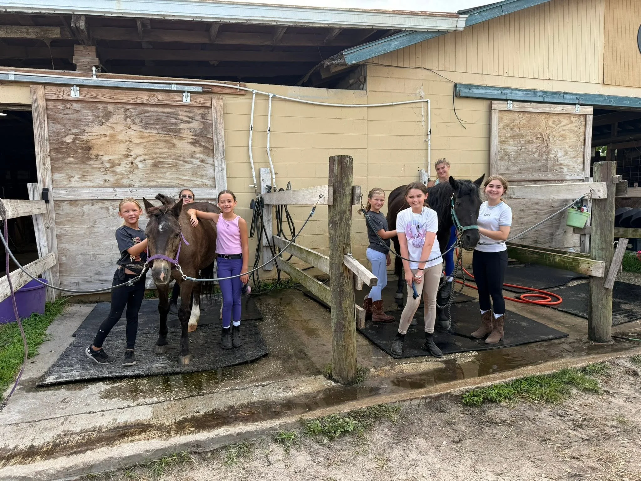 A group of six young girls standing with two horses outside a wooden barn, smiling and posing for the photo.