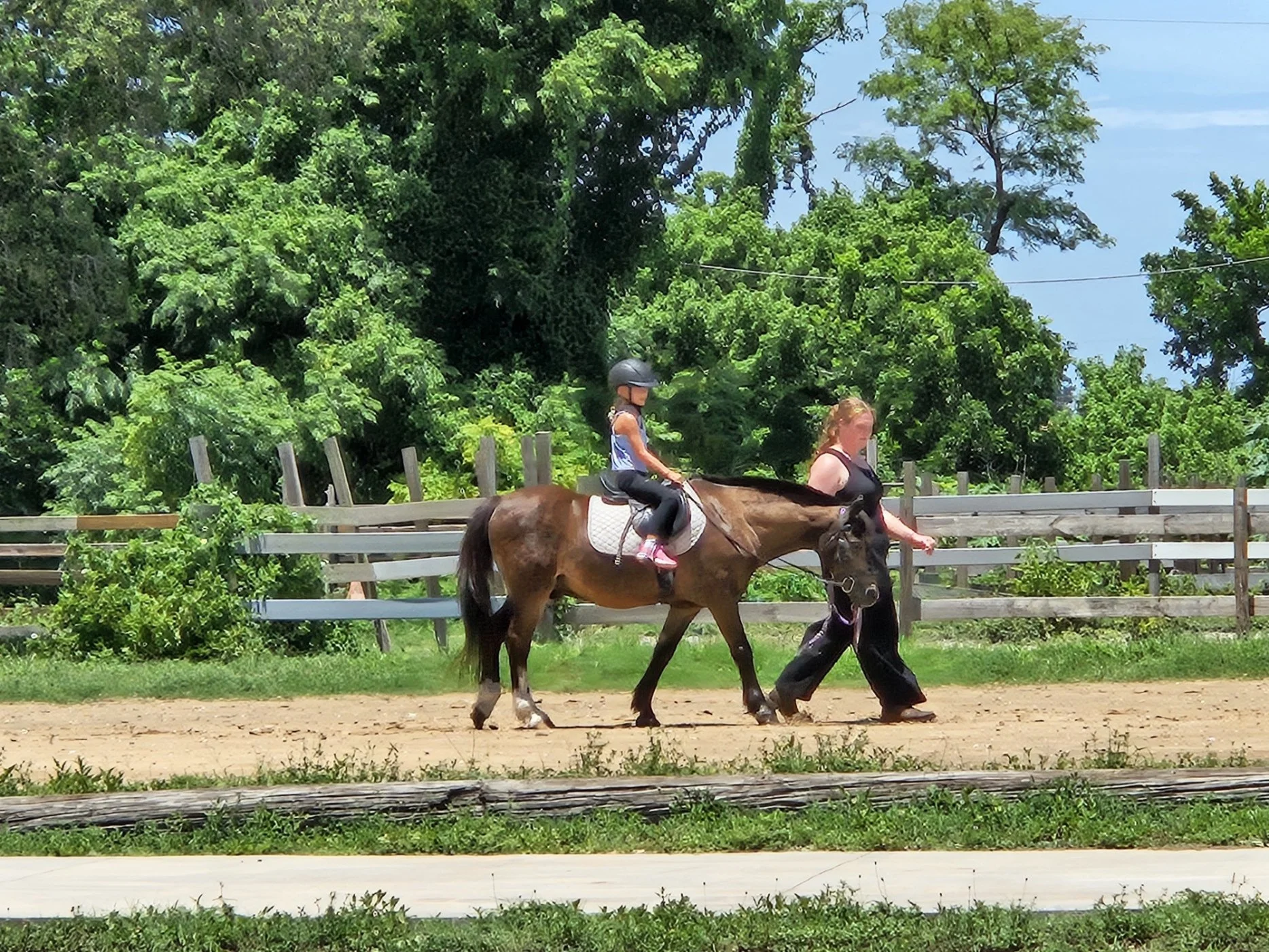 A young girl riding a horse in an outdoor riding arena, accompanied by an adult leading the horse, with lush green trees and a wooden fence in the background on a sunny day.