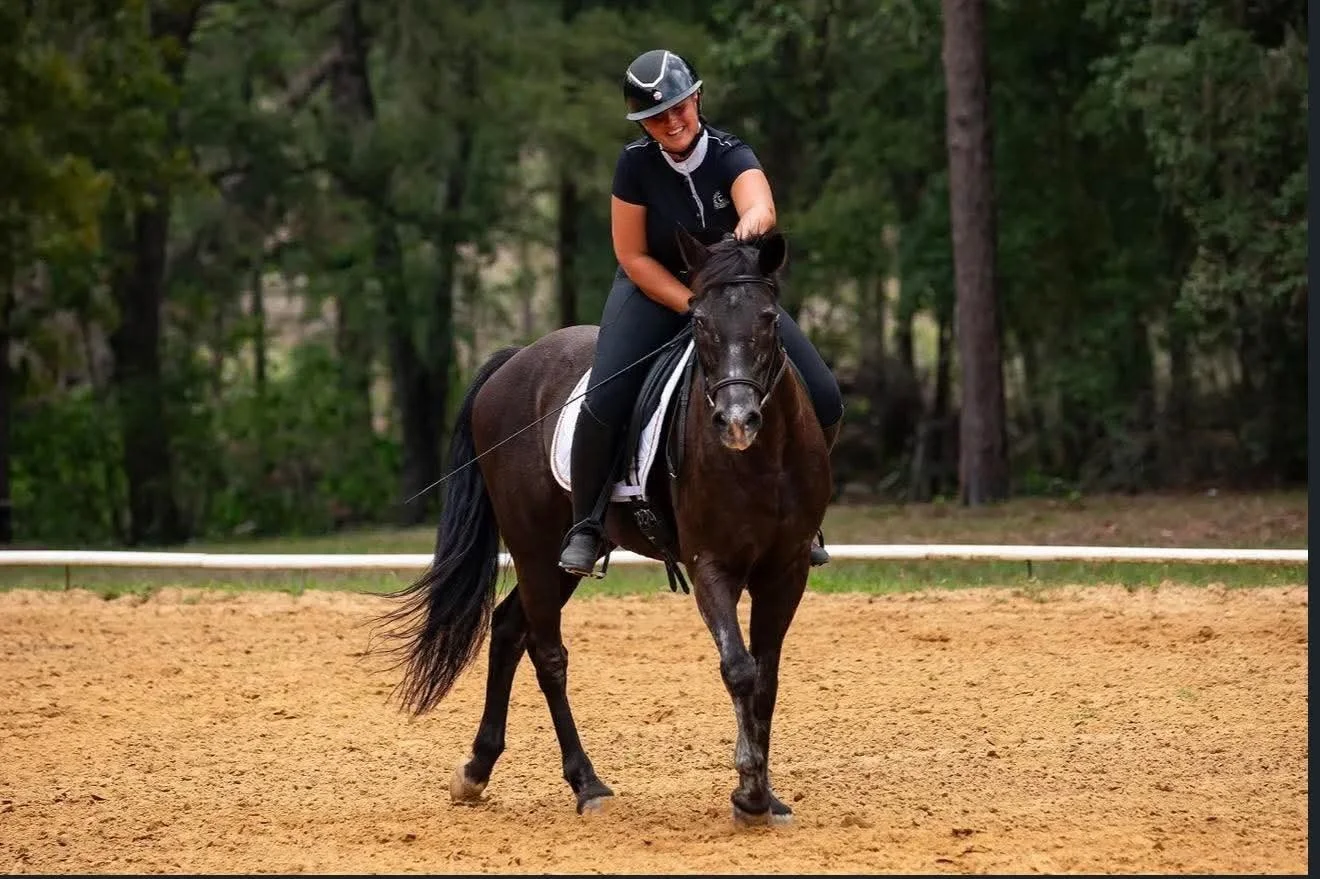 A woman riding a black horse on a dirt arena surrounded by trees during daytime.