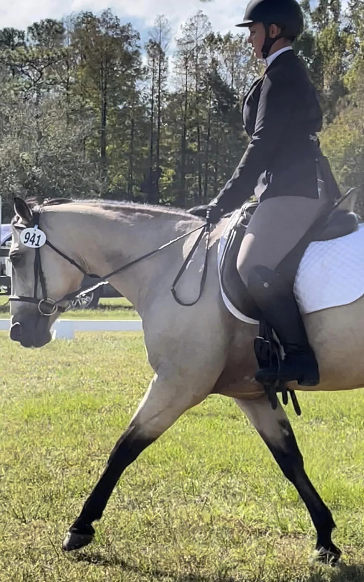 A woman riding a gray horse during a daytime outdoor event, wearing equestrian attire and a helmet, with trees and a blue sky in the background.