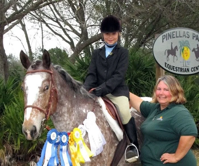 A young girl in equestrian riding attire sitting on a decorated horse, with an adult woman standing beside her, all outdoors with trees in the background and a Pinellas Park Equestrian Center sign.