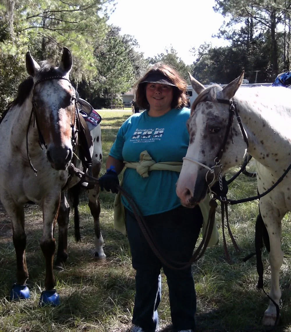 A woman standing outdoors with two spotted horses during daytime, surrounded by trees and grass.