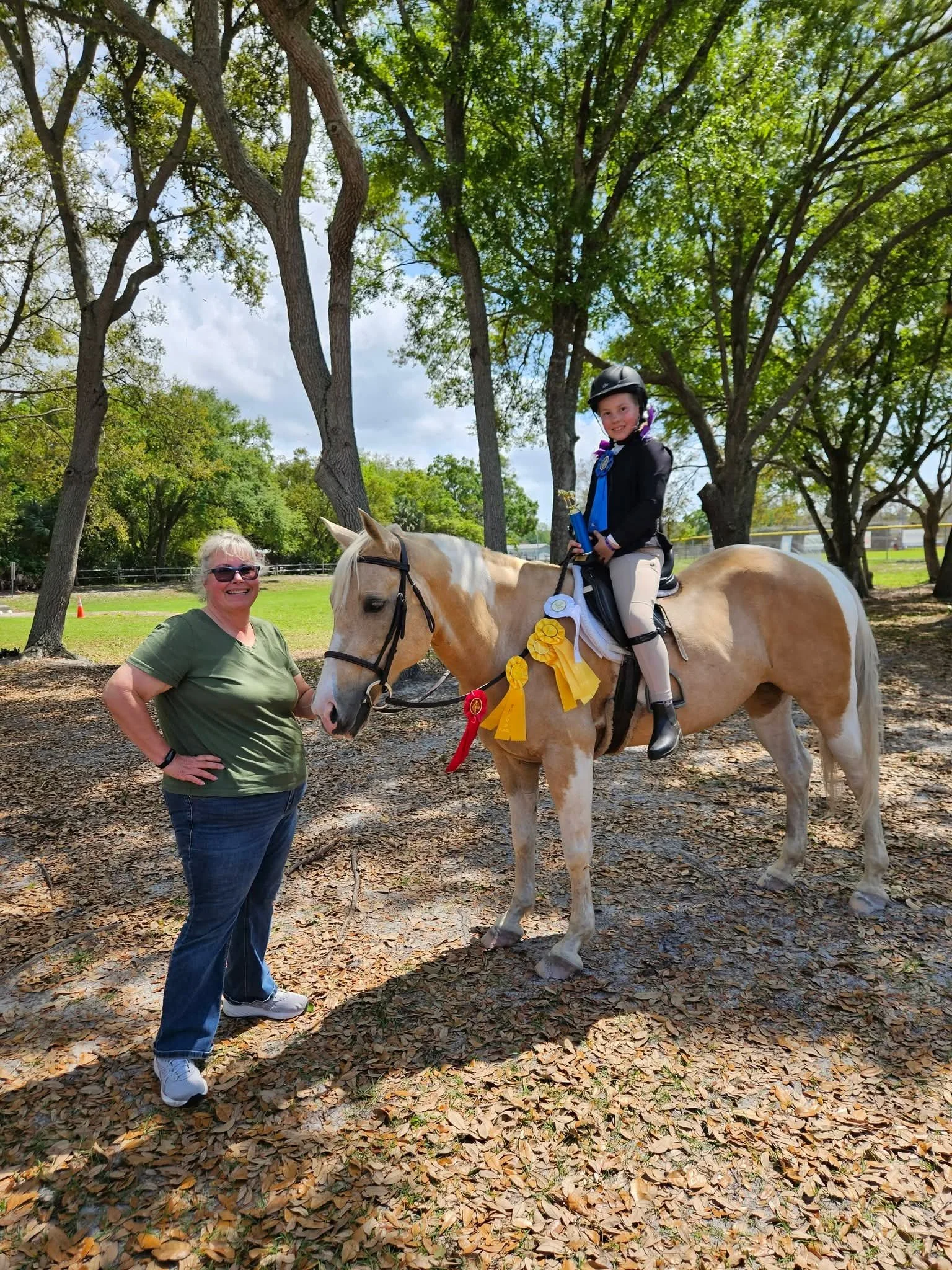 Young girl in equestrian riding gear sitting on a palomino horse with ribbons, next to a woman standing on leaf-covered ground in a park area with tall trees and blue sky.