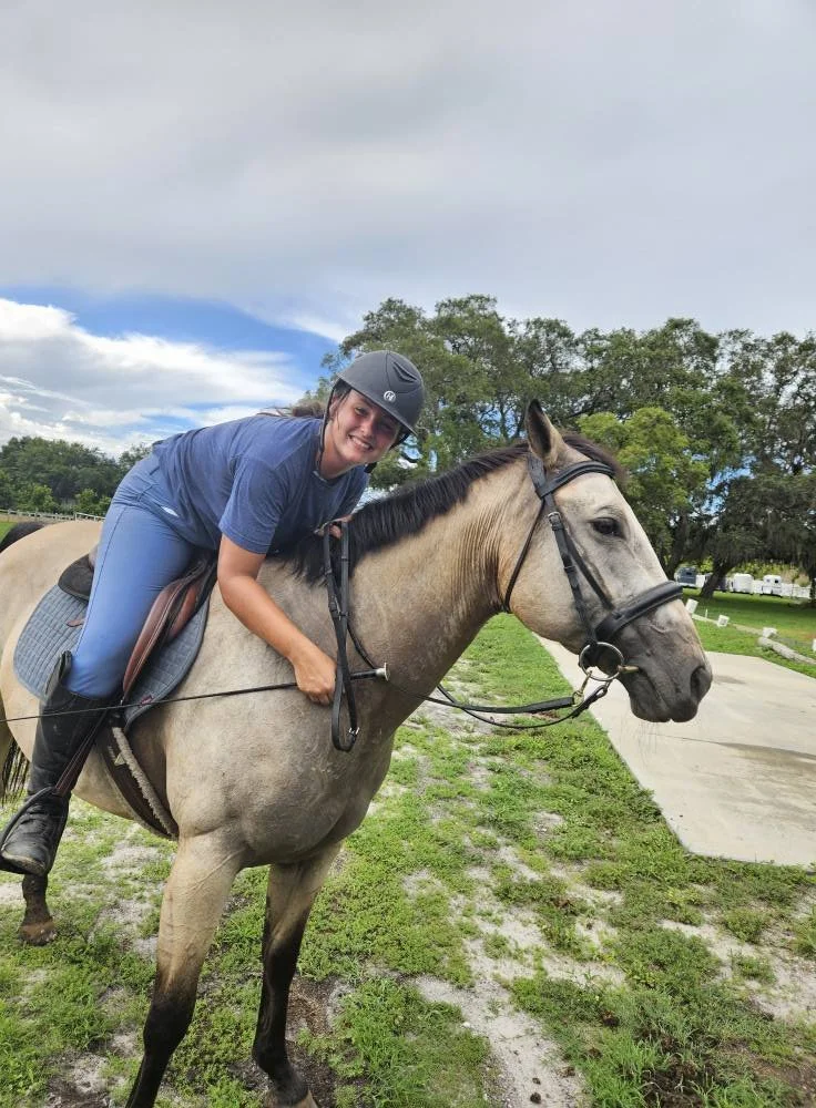 A woman wearing a helmet and blue riding attire riding a light gray horse outdoors on a cloudy day with green trees and grass in the background.