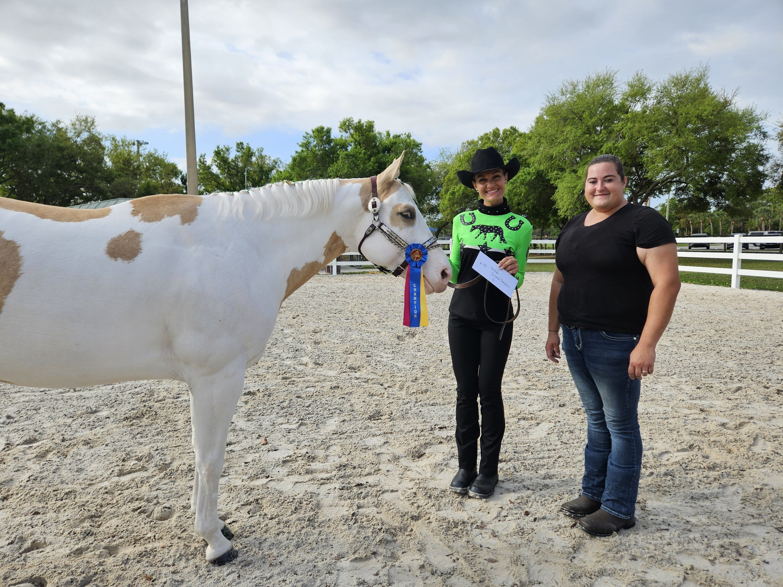 A woman in a black cowboy hat and green rodeo shirt holding a blue ribbon stands beside a white and tan paint horse, with a man in a black t-shirt standing next to her. They are outdoors in a sandy arena with trees and a white fence in the background.