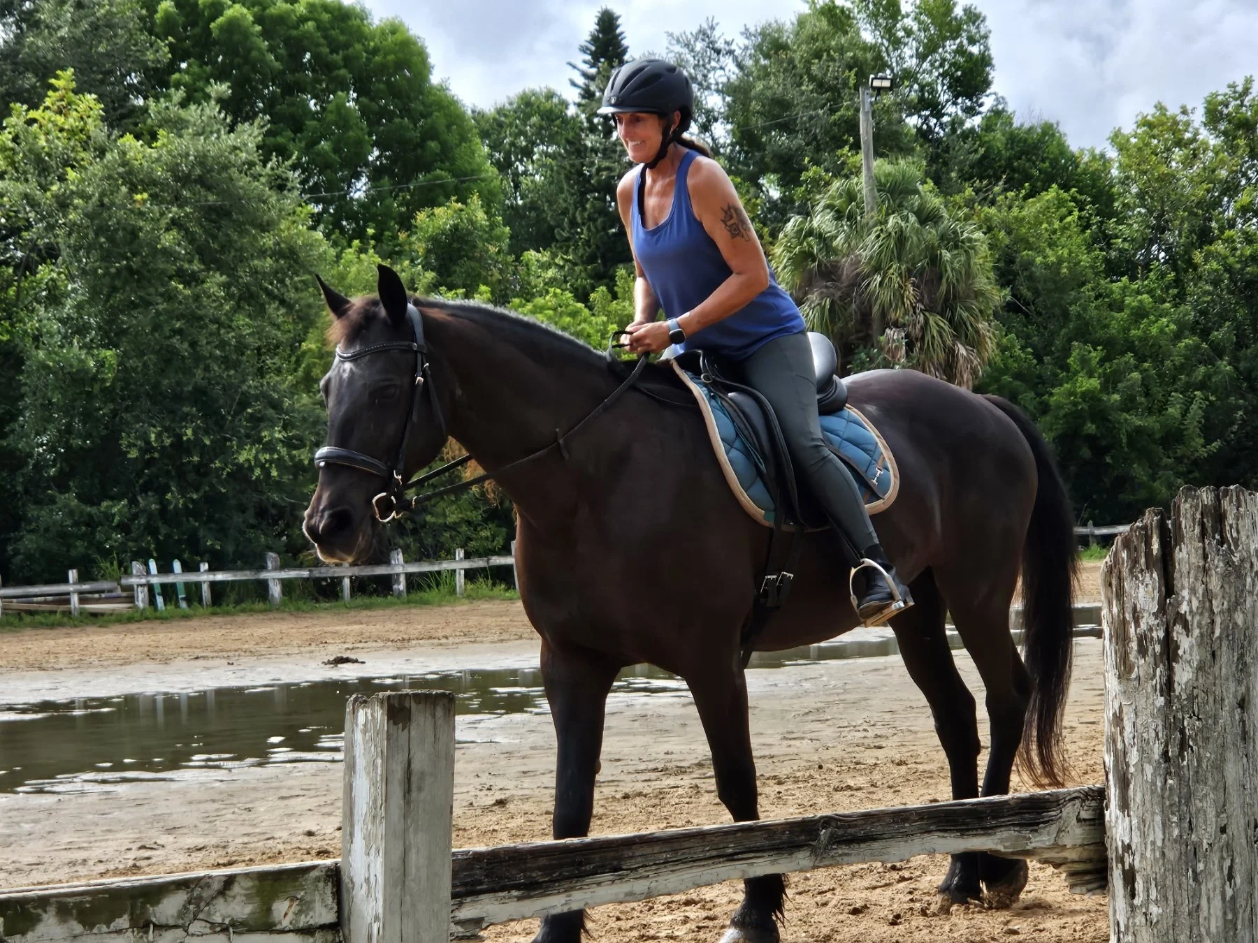 A woman wearing a helmet rides a dark horse on a sandy riding ring. She is smiling and dressed in a blue tank top and black riding pants, with a tattoo on her upper arm. There are trees and a cloudy sky in the background.