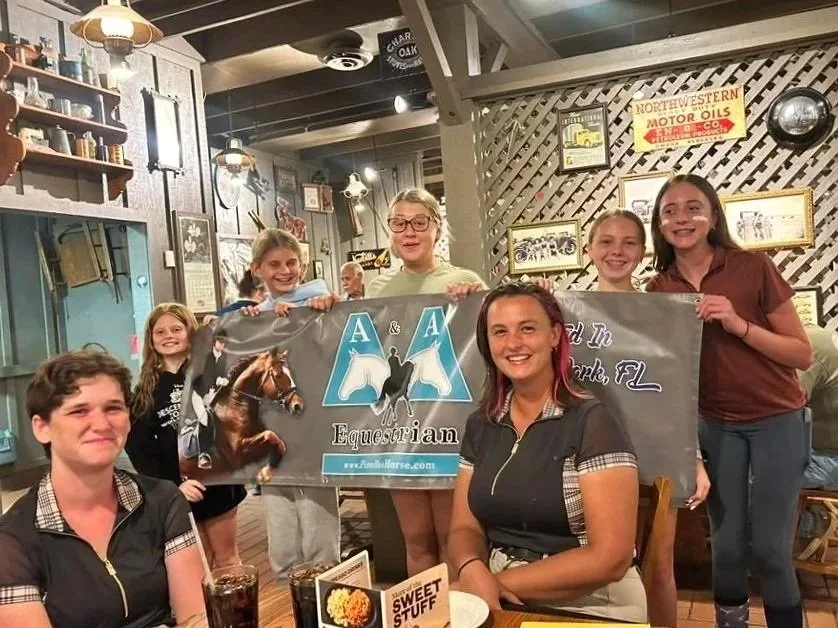 Group of girls and women holding an equestrian banner inside a restaurant or cafe with vintage decor.