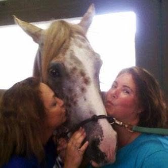 Two women kissing a white horse with brown spots on its face.