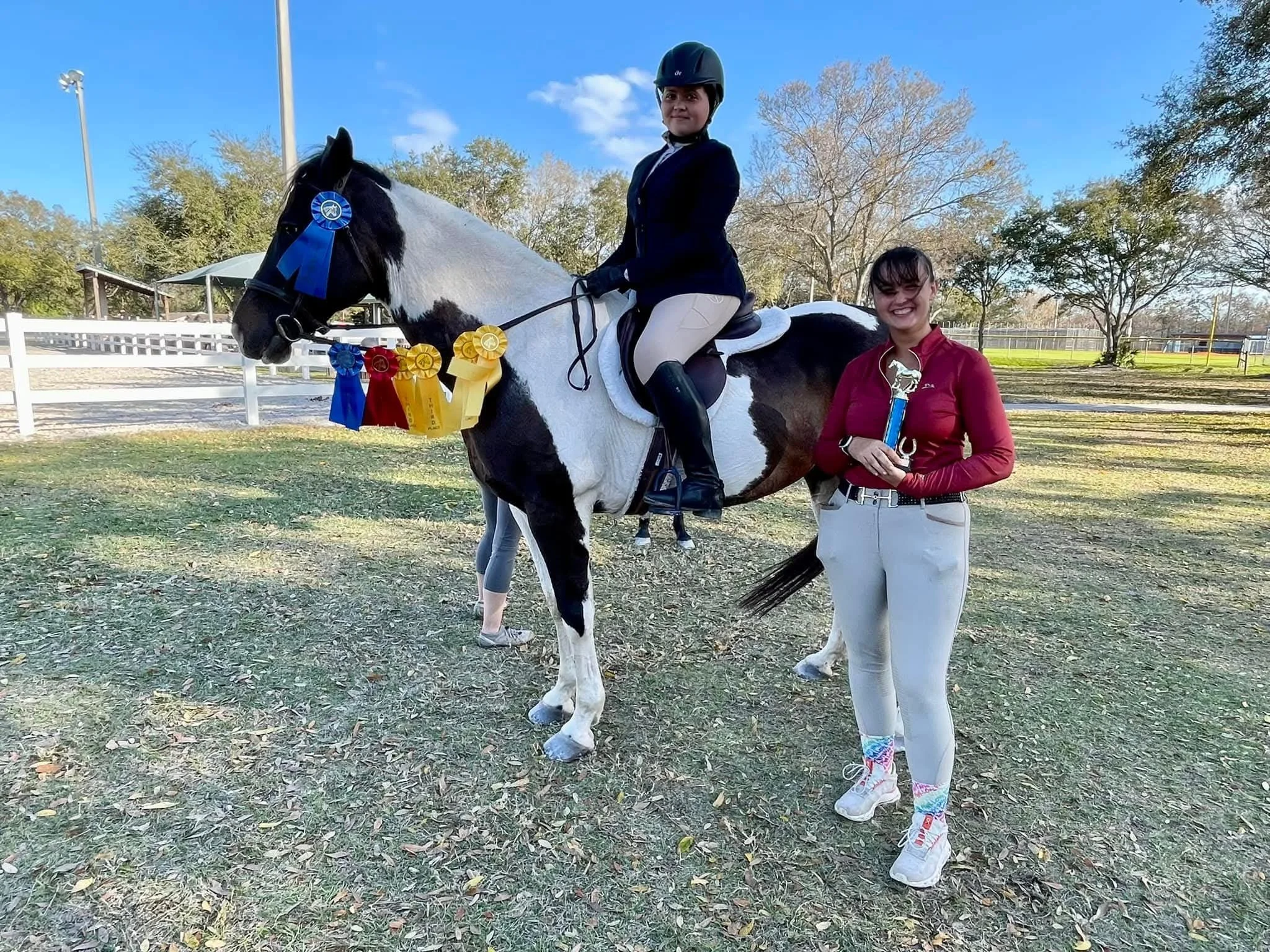 A young woman sitting on a black and white horse with ribbons attached to its bridle, while another woman stands beside them holding a trophy. They are outdoors in a fenced area with trees and a clear sky.
