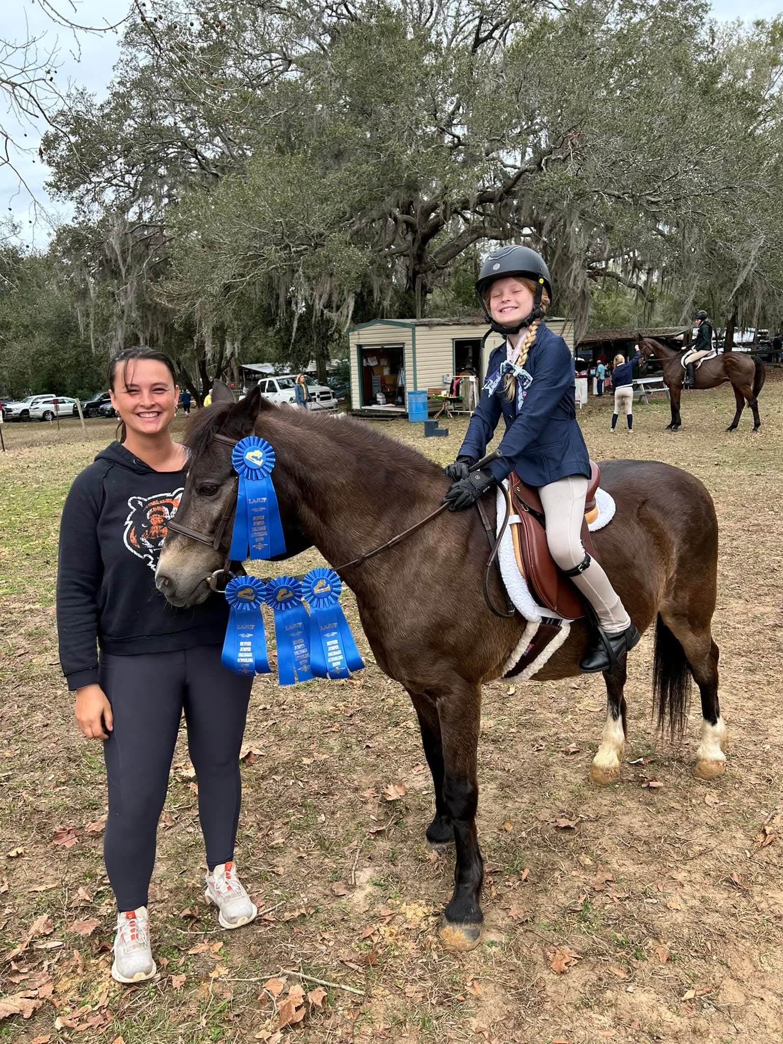 A girl riding a brown horse with a blue ribbon, standing on grass with a woman beside her. The girl is wearing a helmet, jacket, and riding pants, smiling with closed eyes. The woman is smiling, standing next to the horse, wearing a black hoodie and 