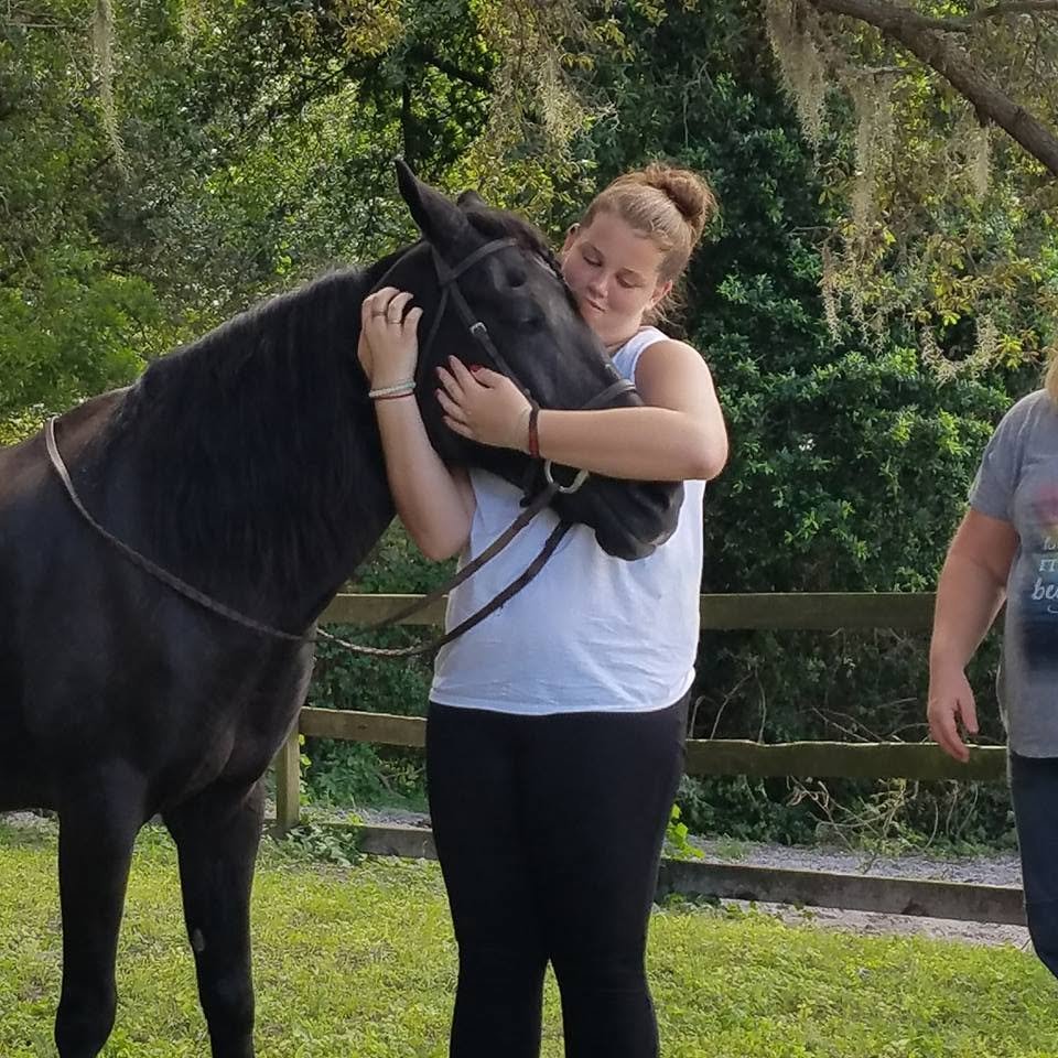 A young woman hugging a black horse outdoors in a grassy area with trees and a wooden fence in the background.