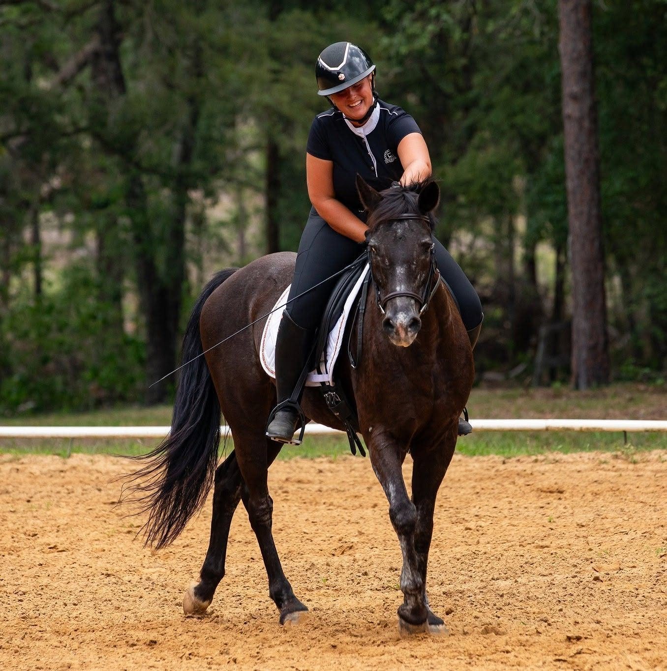 A woman riding a dark brown horse on a dirt track in a wooded area, smiling and wearing a helmet and black riding clothes.
