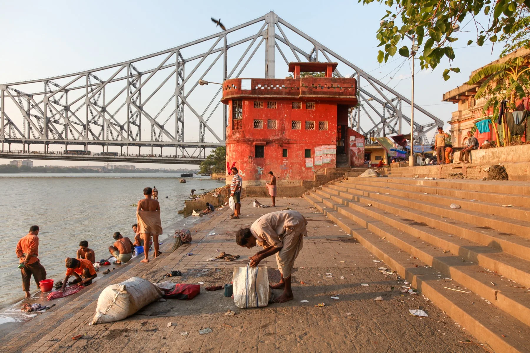 People gather along the steps of the Hooghly River at sunset in Kolkata, West Bengal, India. Bathing, washing and quiet moments mark everyday life beneath the iconic Howrah Bridge on the city’s historic waterfront.