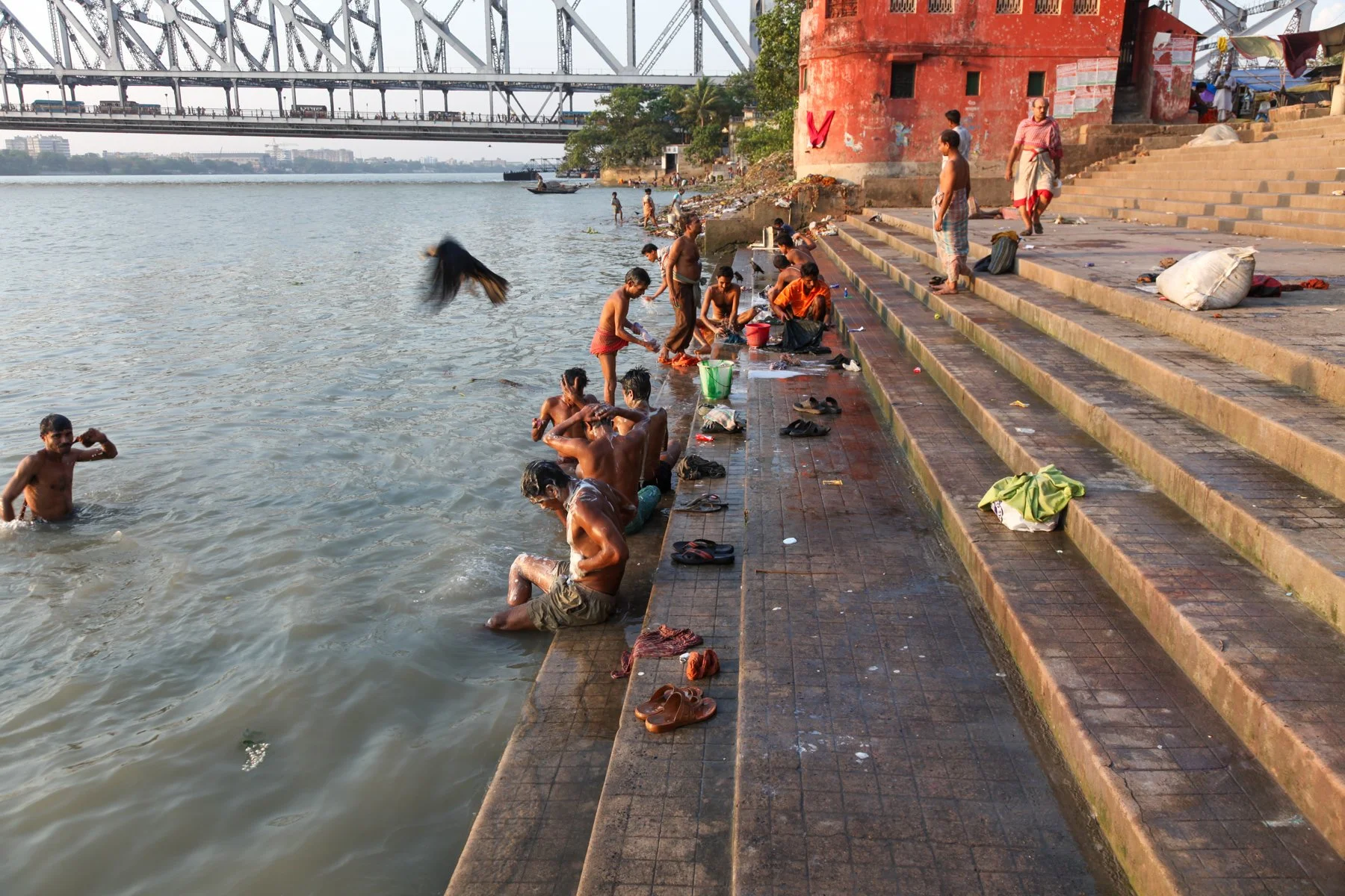 People bathe and wash at a riverside ghat on the Hooghly River beneath Howrah Bridge in Kolkata, West Bengal, India. The evening light and everyday rituals reflect the close relationship between the city and its river.
