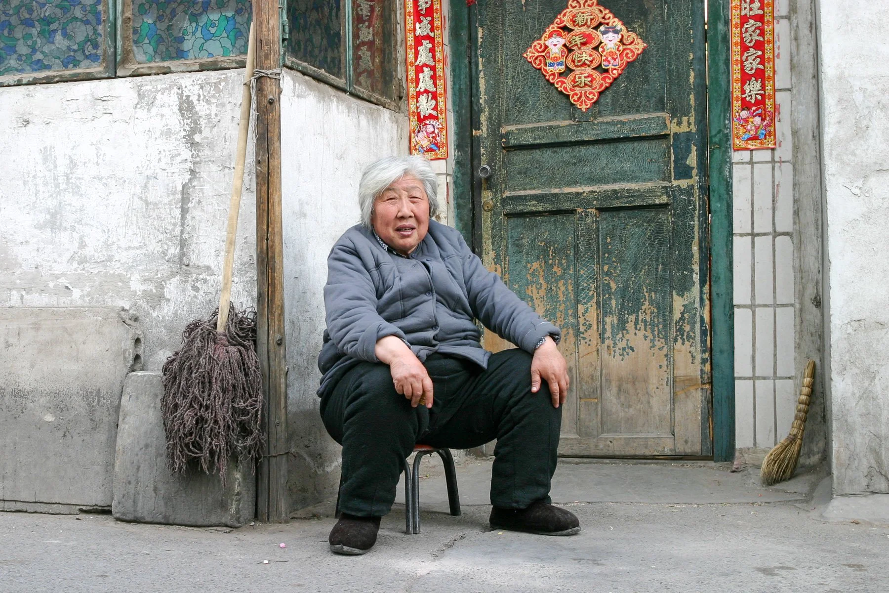 An elderly woman sits on a small stool outside her home in a hutong neighborhood in Beijing, China, looking directly at the camera.Documentary street photograph taken in 2005.