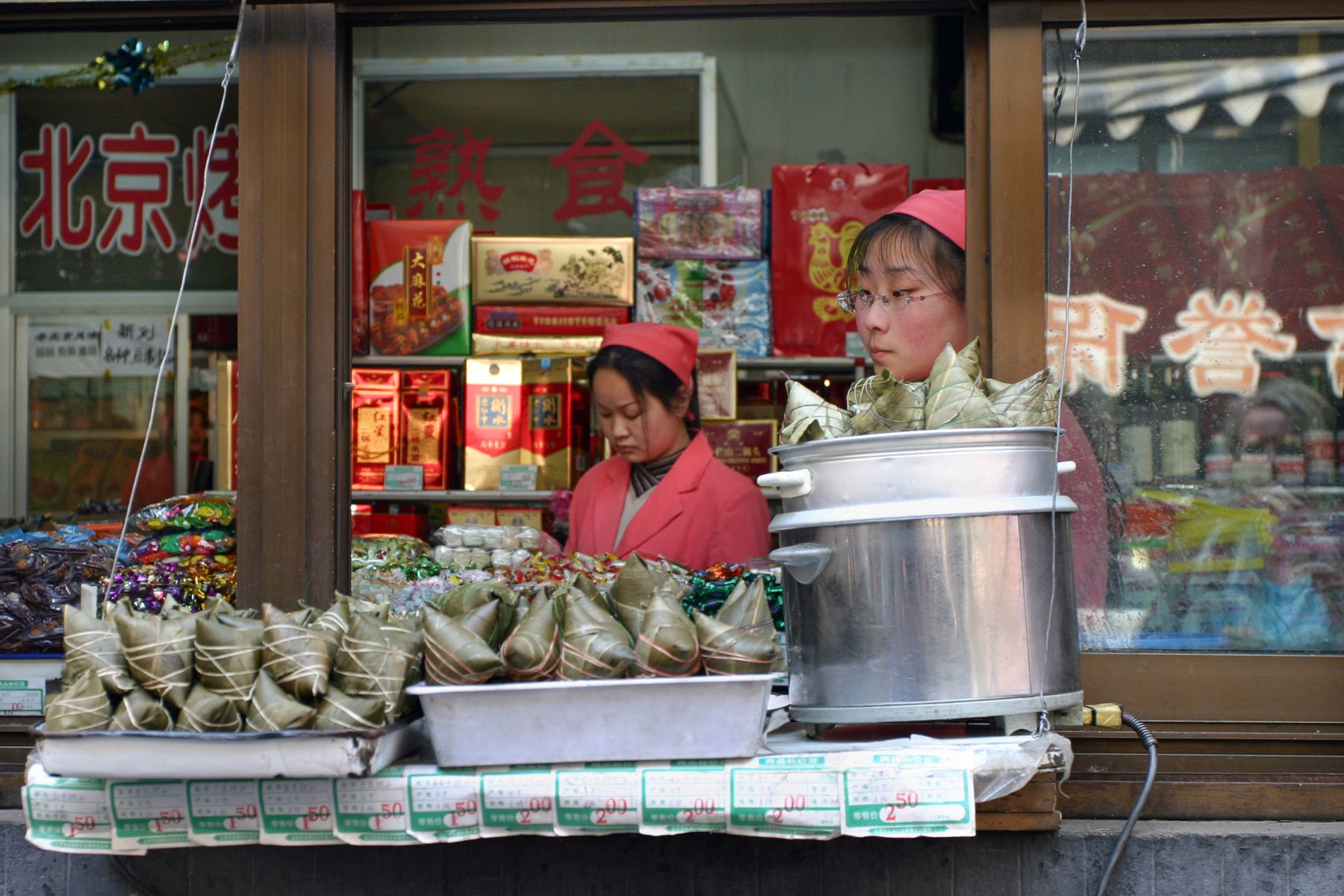 Two women wearing red caps work behind the open serving window of a small food stall in Beijing, China.Documentary photograph taken in 2005.