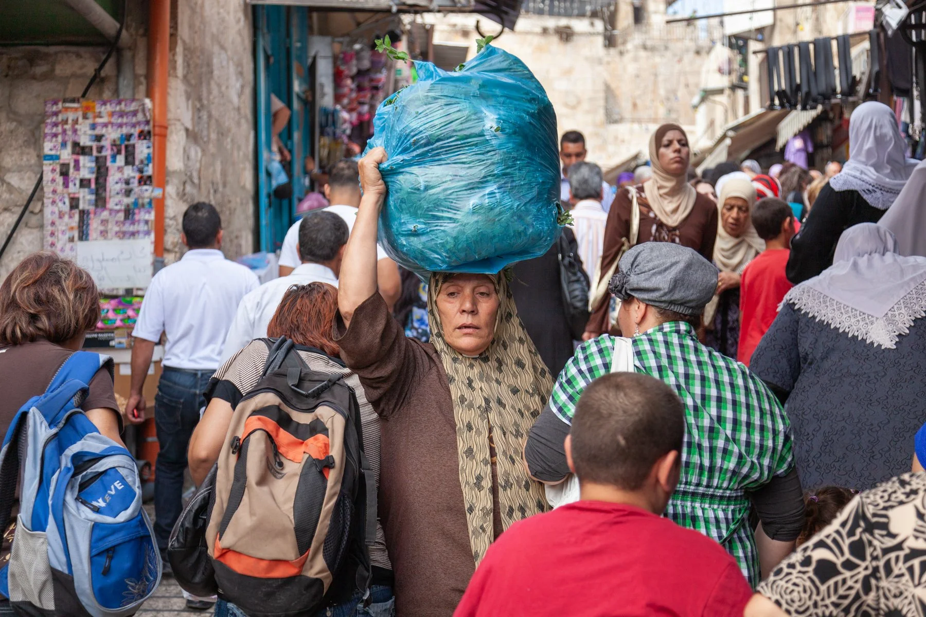 Woman carrying a large blue sack on her head through a crowded market in Jerusalem’s Old City.