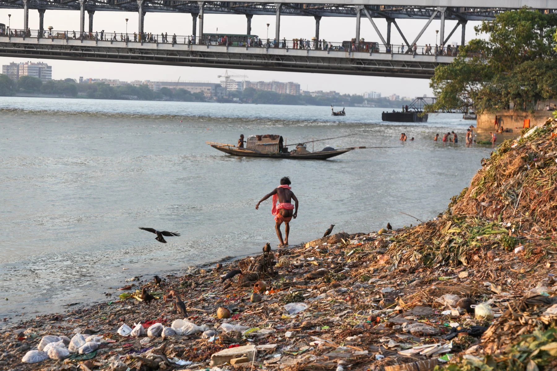 A boy walks along the littered banks of the Hooghly River beneath Howrah Bridge (Rabindra Setu) in Kolkata, West Bengal, India. The scene contrasts everyday life, urban pollution and one of the city’s best-known landmarks.
