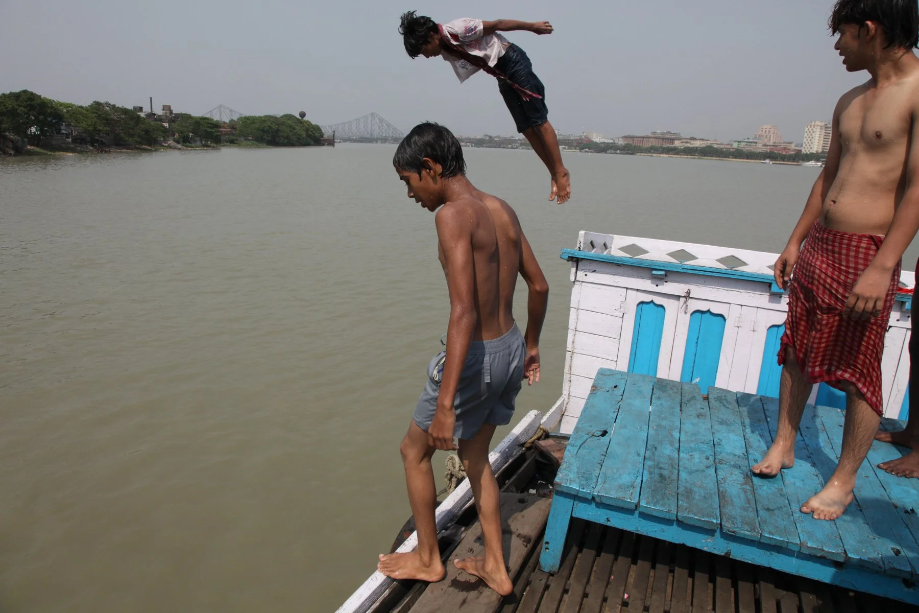 A boy dives from the deck of a ferry into the Hooghly River while friends watch on a hot day in Kolkata, West Bengal, India. Swimming and diving from ferries and riverbanks are a familiar part of everyday life along the Hooghly. The Howrah Bridge is 