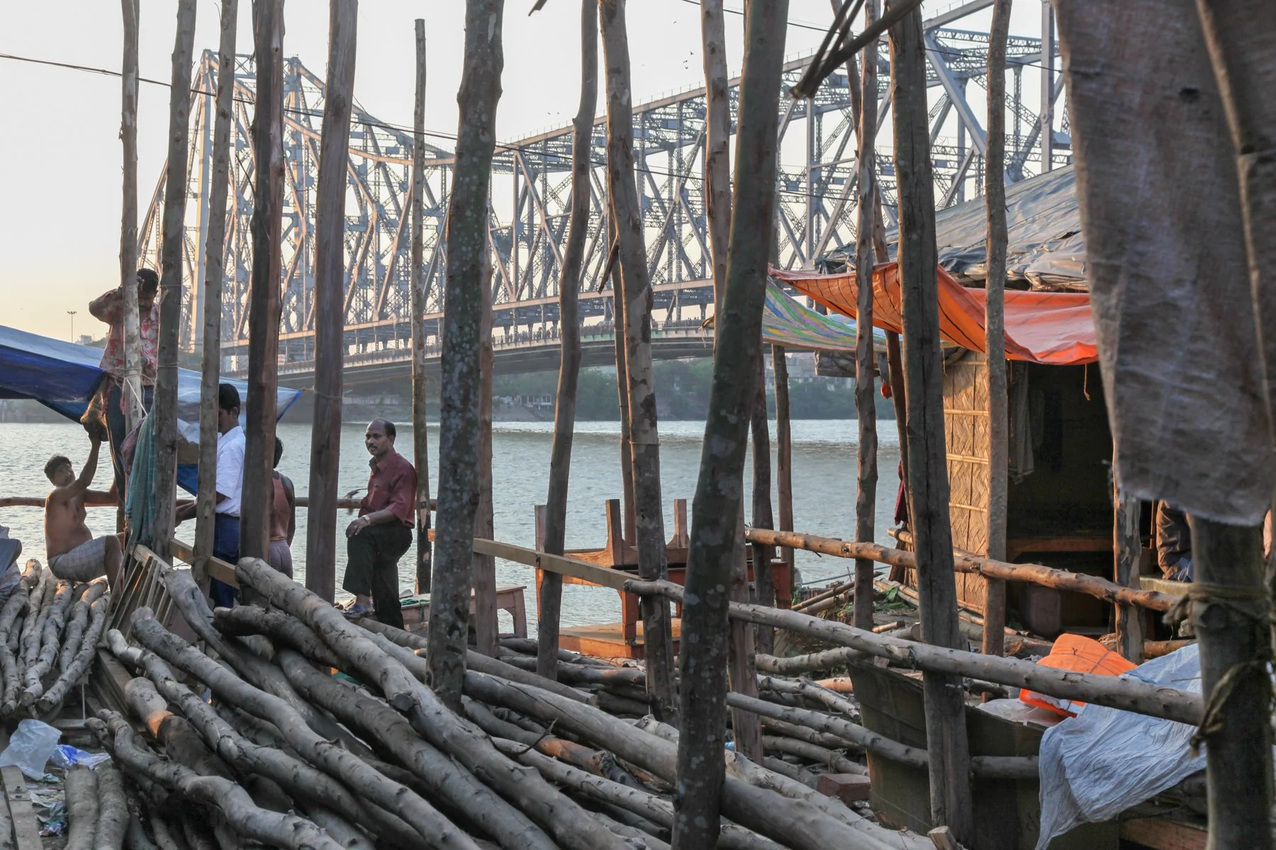 Wooden poles, boats and temporary riverside shelters stand along the Hooghly River with Howrah Bridge in the background, Kolkata, West Bengal, India.