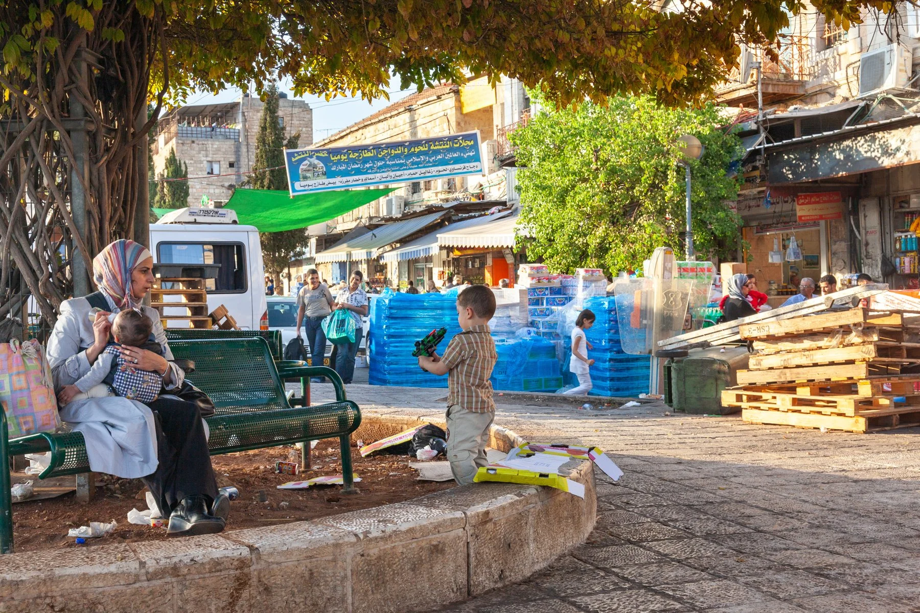 Mother holding her child on a bench while a young boy plays nearby in Jerusalem.