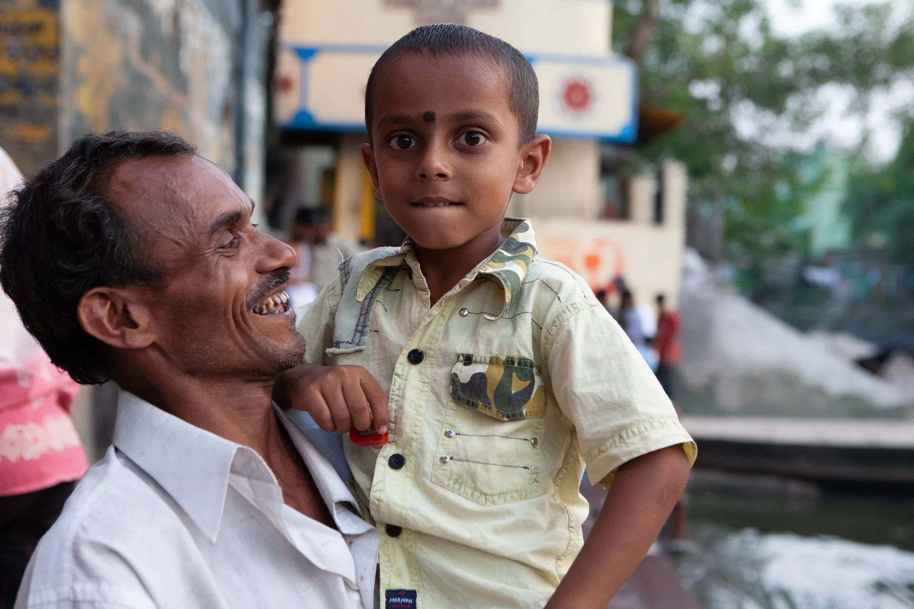 A father smiles while holding his young son beside the Hooghly River in Kolkata, West Bengal, India. The portrait captures a quiet family moment along the riverbank, where everyday life unfolds around one of the city’s most important public spaces.