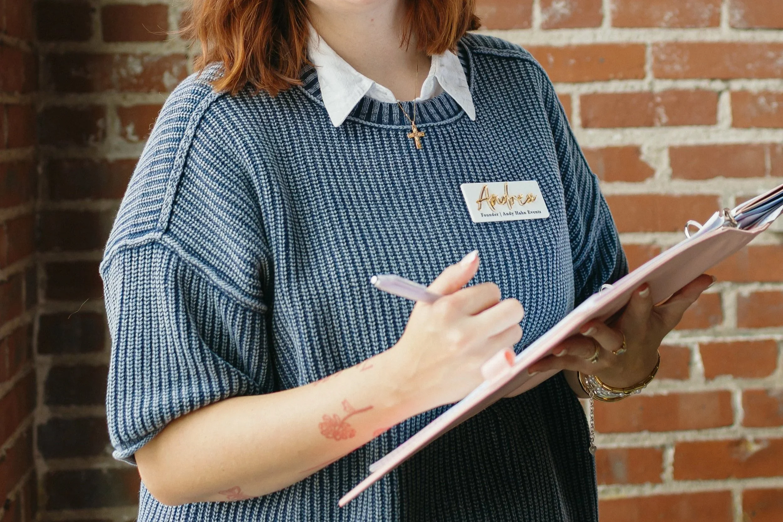 A woman with red hair wearing a blue and white striped sweater, a white collared shirt, and gold jewelry, holding a pink notebook and pen in front of a brick wall.