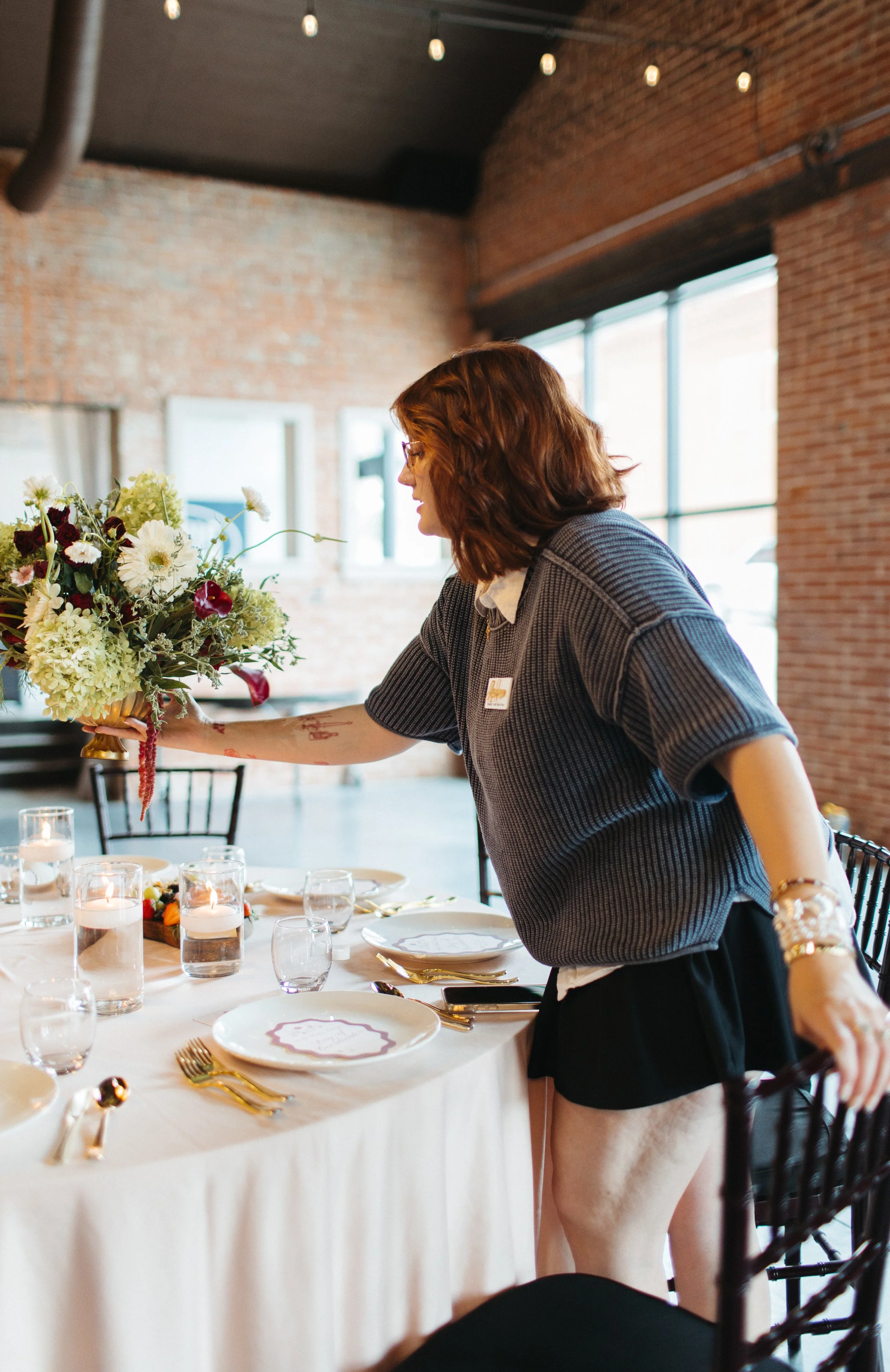 A woman with red hair and glasses is decorating a table with a floral centerpiece at a festive event. The table has white tablecloths, plates, gold utensils, and candles. The background features brick walls, large windows, and hanging string lights.