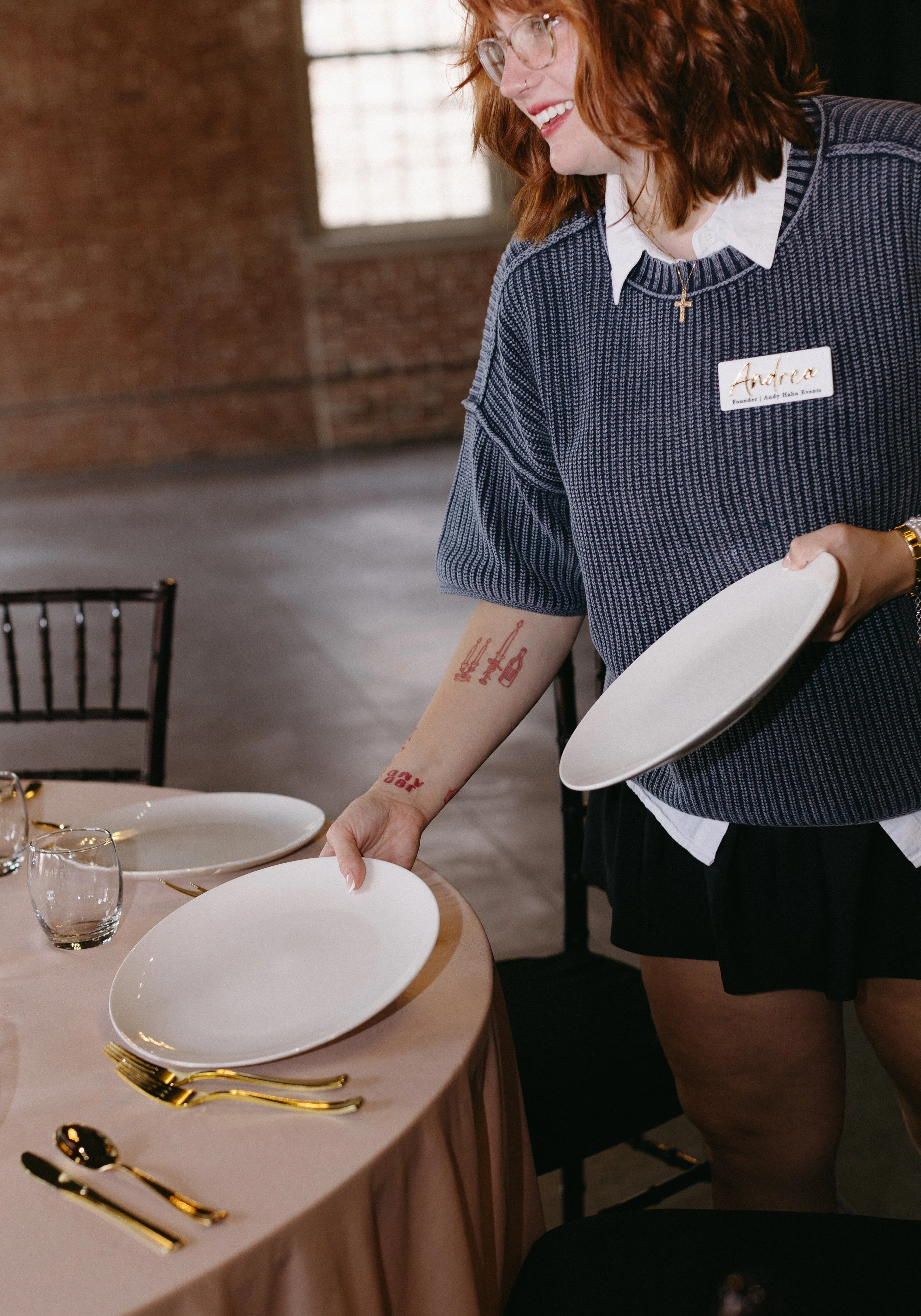 A woman with red hair, glasses, and a necklace with a cross, is setting a dinner table with white plates, gold utensils, and glasses, in a warmly lit room with brick walls and large windows.