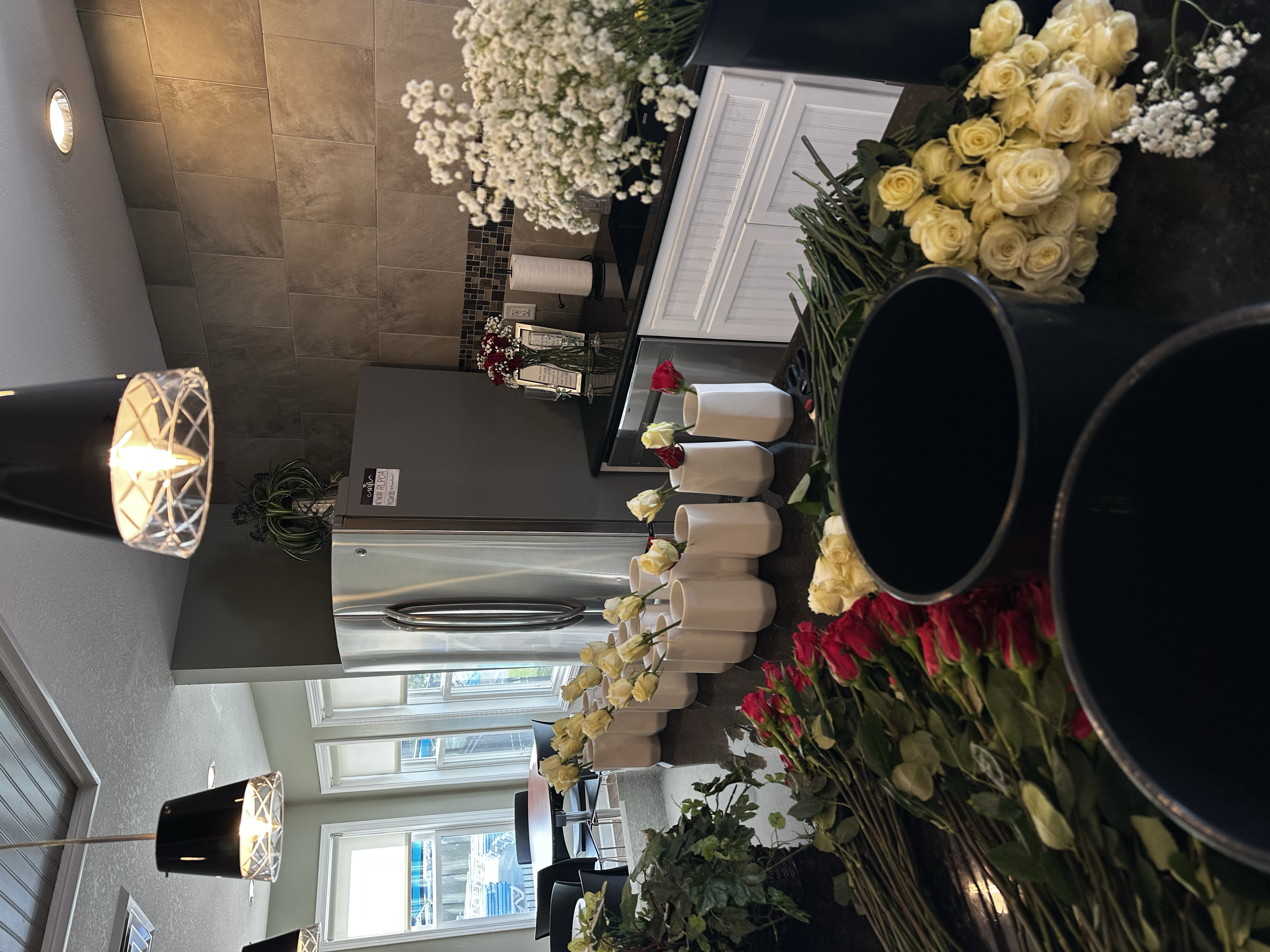 A room with a window, decorated wall, and a table filled with various flowers, including yellow roses, white roses, and small white blossoms, arranged in vases and containers.