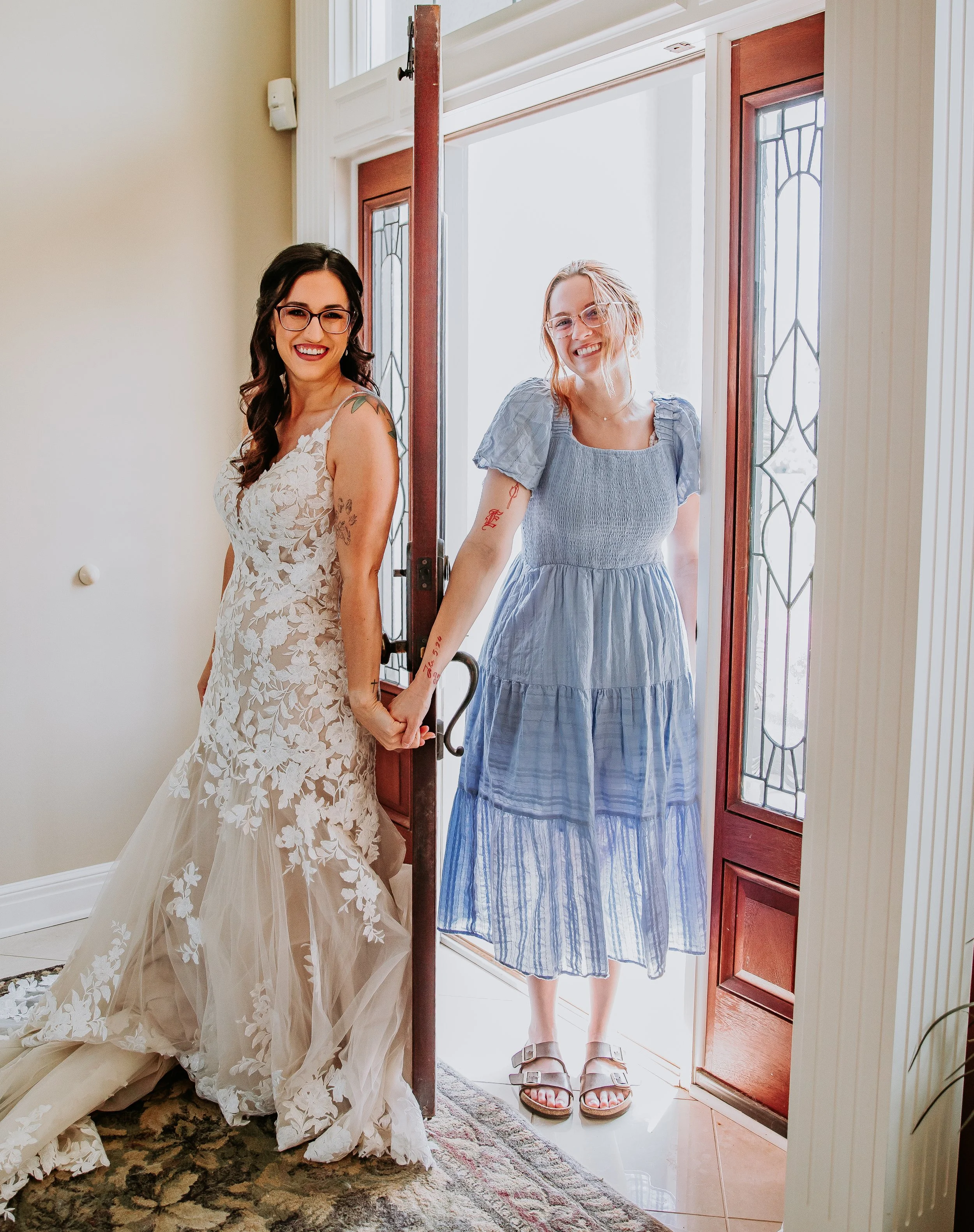A bride in a floral lace wedding dress standing inside near the door, holding hands with a woman in a blue striped dress and sandals, who is standing in the doorway.