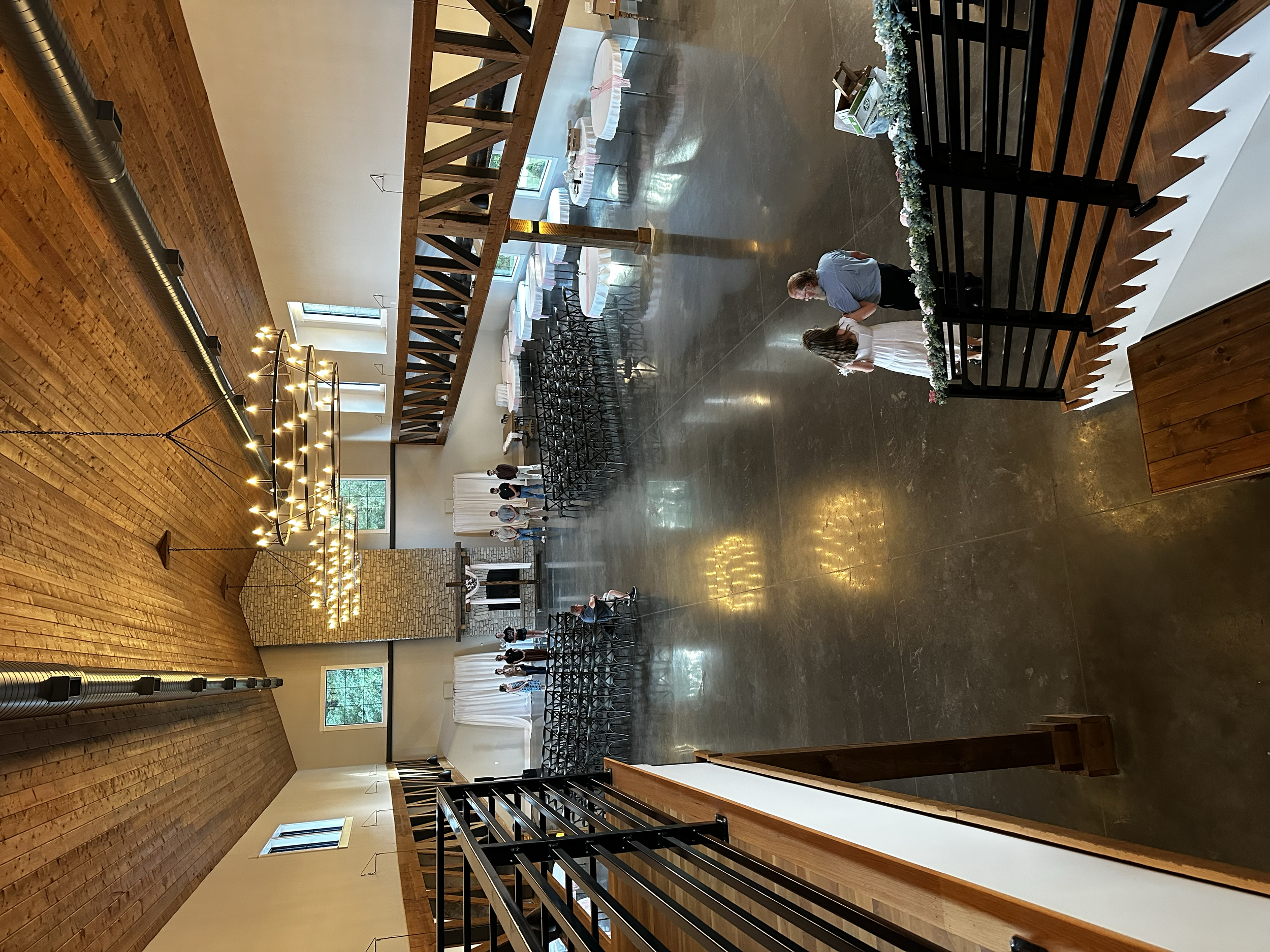 People standing and conversing in a spacious room with wooden walls and high ceilings, decorated with string lights and floral arrangements.