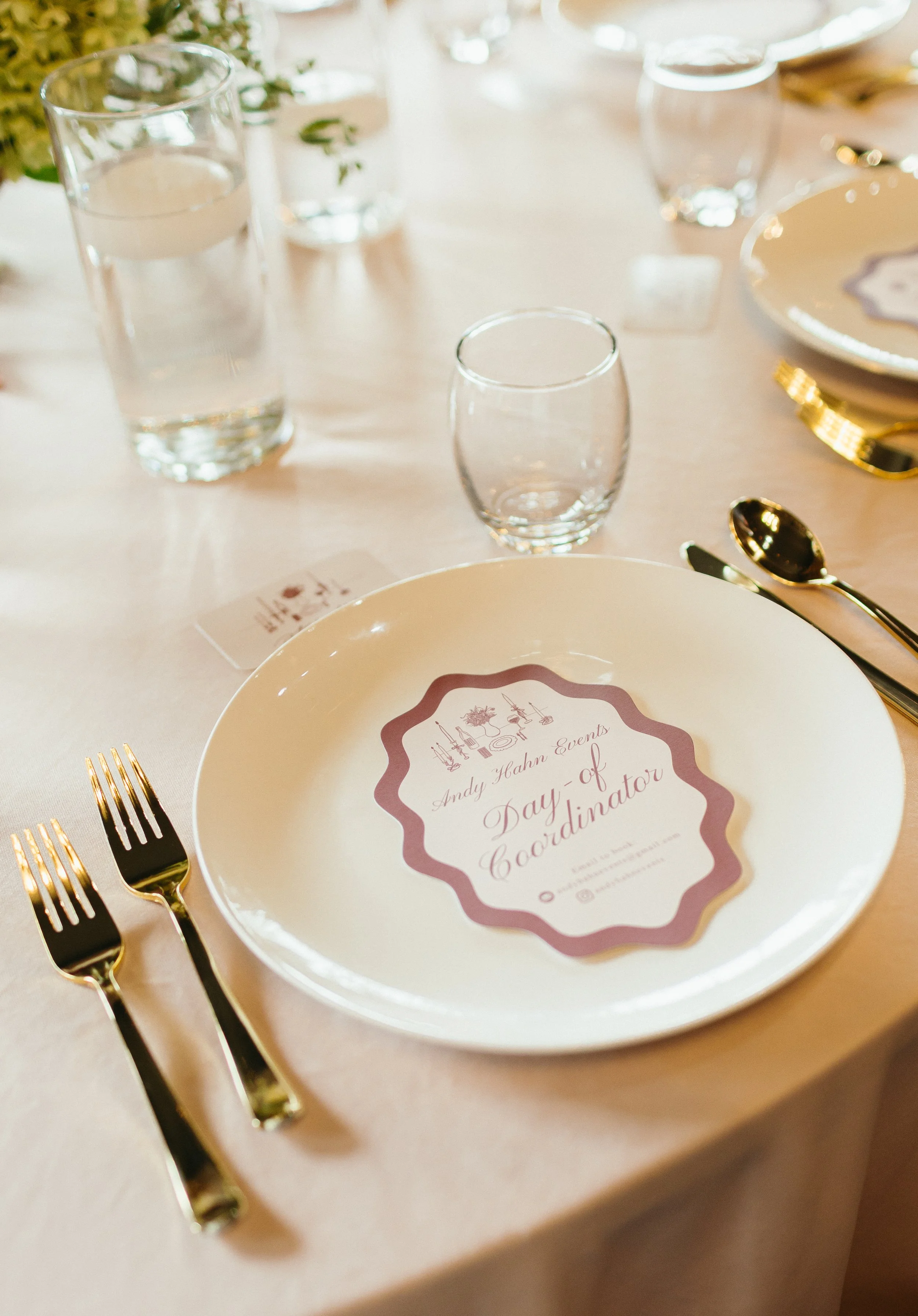 Elegant table setting with a white plate, gold utensils, and a printed paper on the plate that reads 'Day of Coordinator.' Clear water glasses and a menu card with decorative border and cursive font on a beige tablecloth.