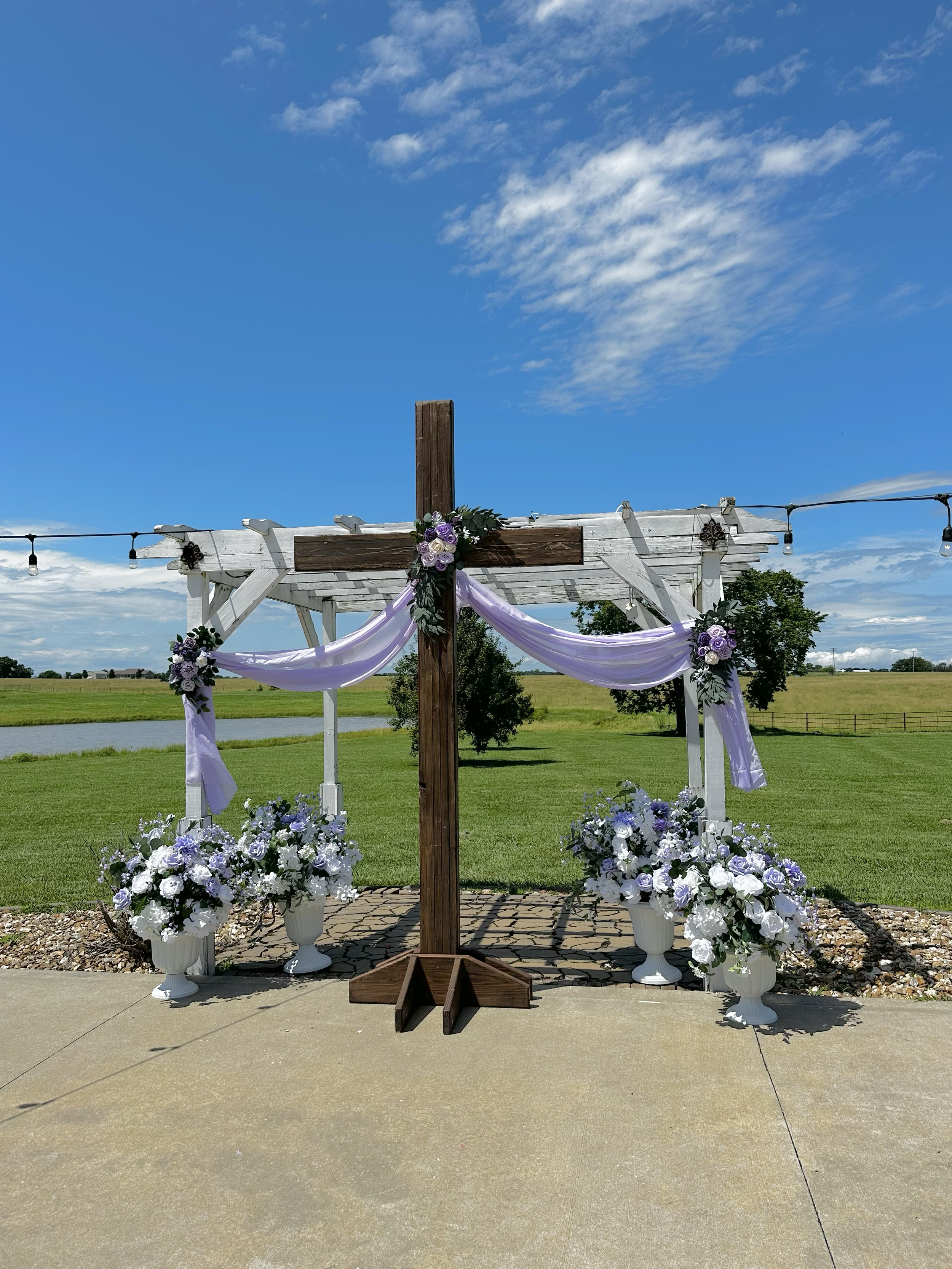 An outdoor wedding altar decorated with purple and white flowers, white drapery, and a wooden cross, set on a paved area with green grass, trees, and a blue sky with some clouds in the background.