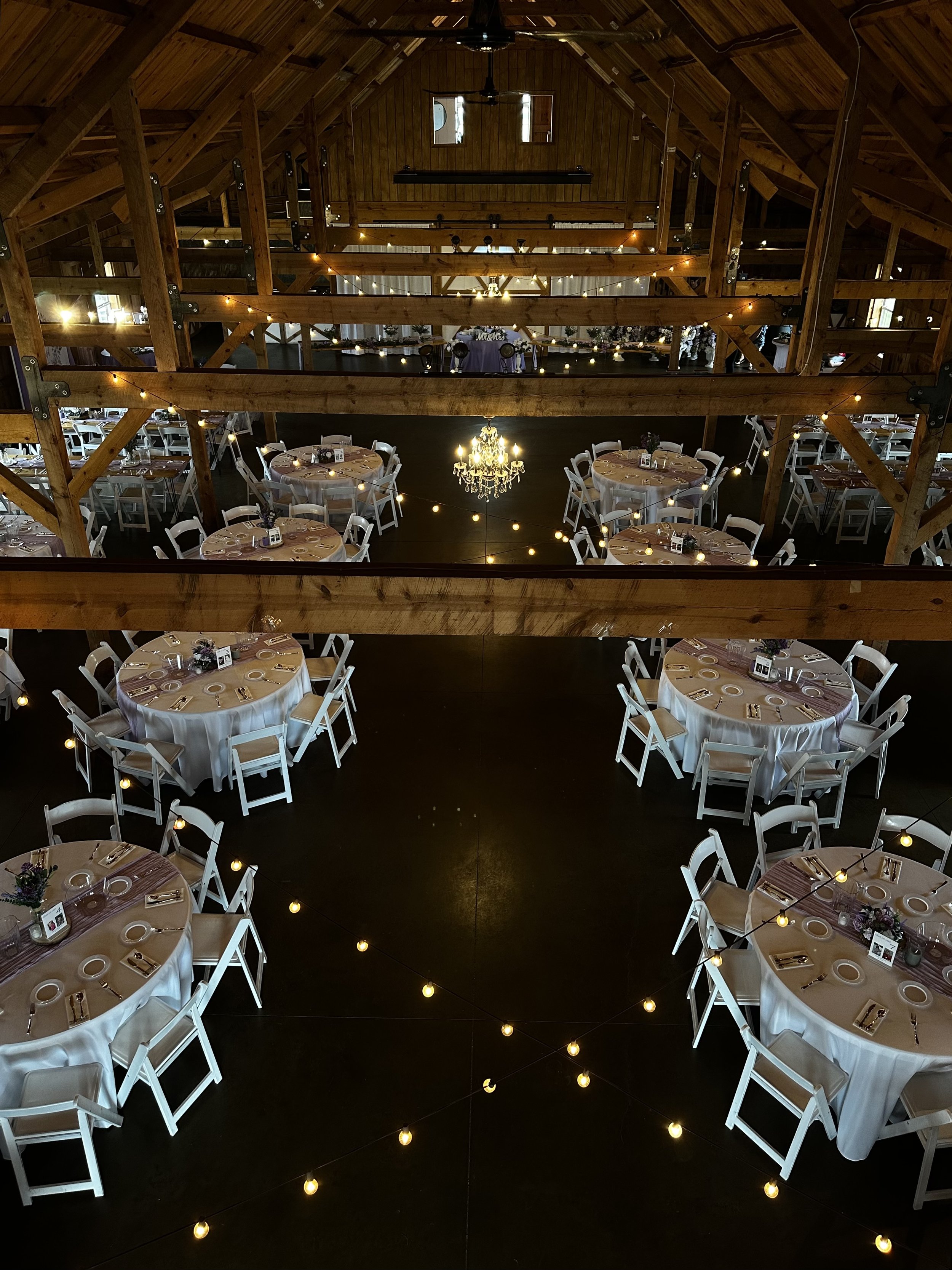 Indoor wedding reception venue with round tables covered in white tablecloths, decorated with purple flowers and candles, arranged around a central chandelier, with wooden beams and string lights overhead.
