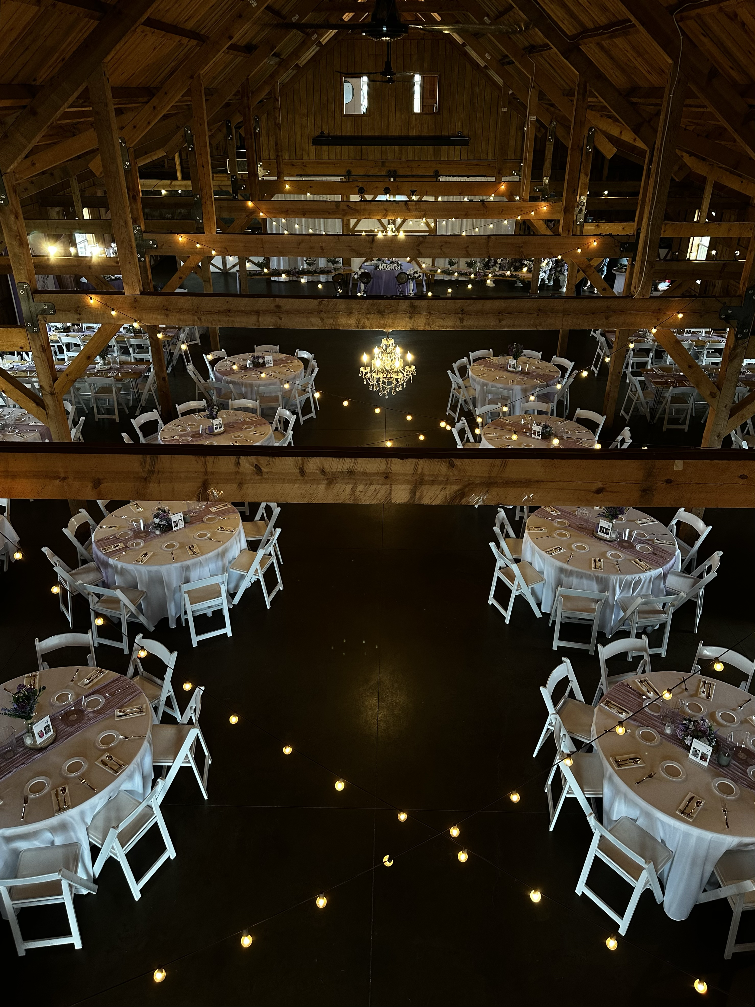 Inside of a rustic wooden barn decorated for a wedding reception with round tables covered in white tablecloths, white chairs, string lights, and a chandelier in the center.