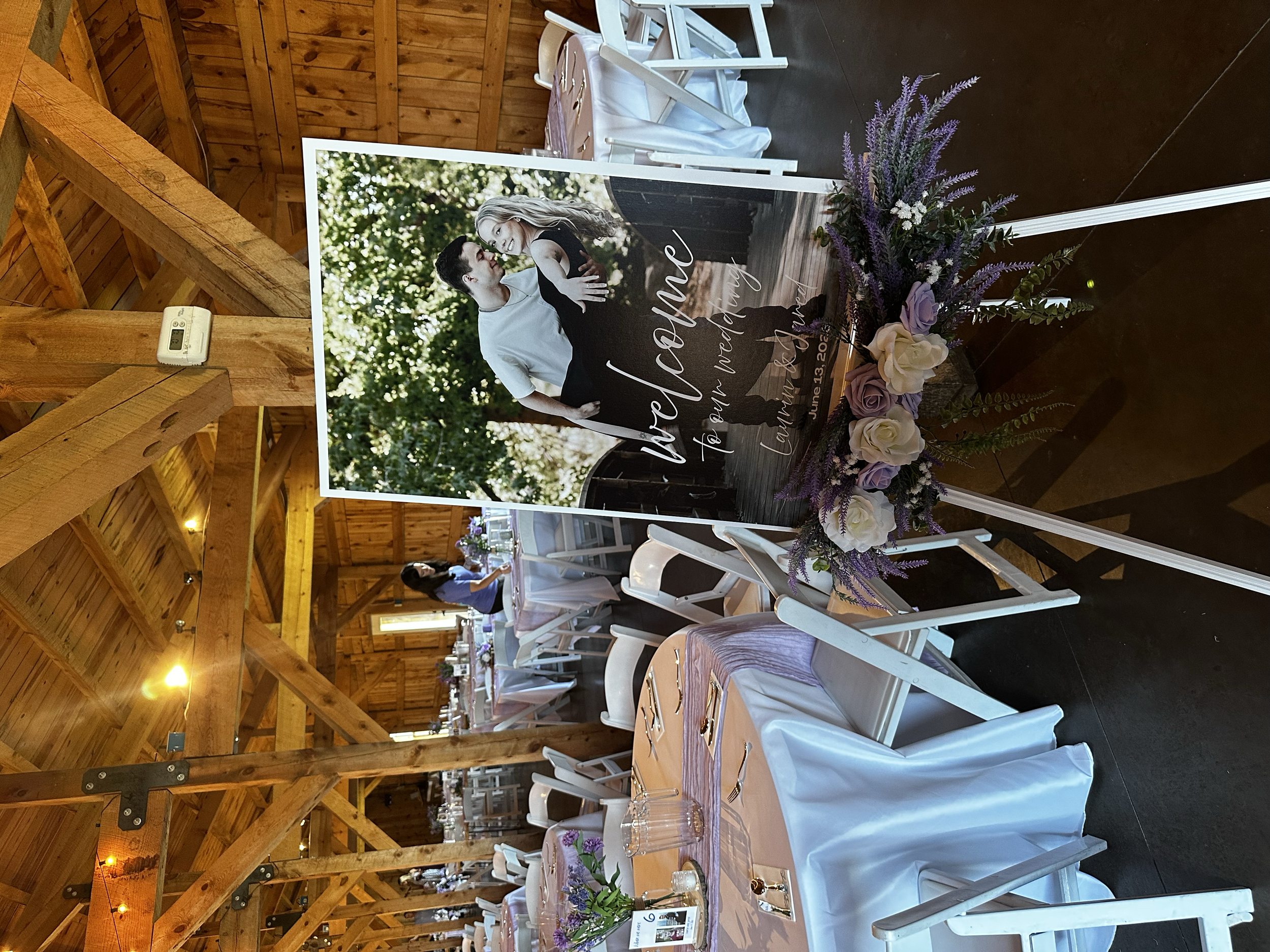 A wedding reception setup with round tables covered in light pink tablecloths, decorated with floral centerpieces featuring purple and white flowers, in a rustic wooden hall with high ceilings and exposed beams. 