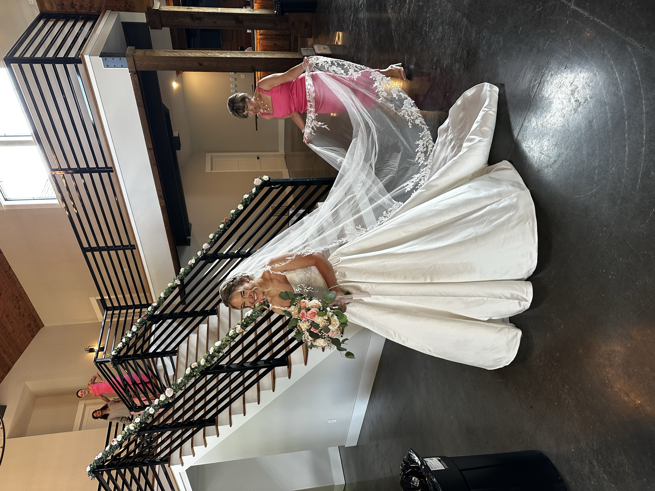 A bride in a white wedding gown holding a bouquet of pink and white roses, smiling, with her veil flowing behind her.