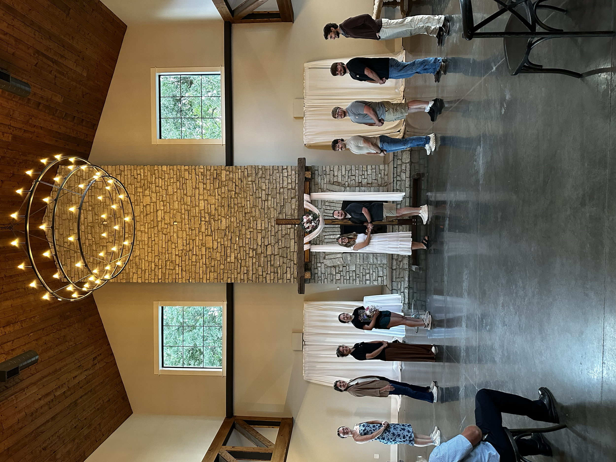 A group of people standing in a line indoors, facing a woman in a white dress with floral decor. The room has a high wooden ceiling, stone brick wall, large windows, and black chairs.