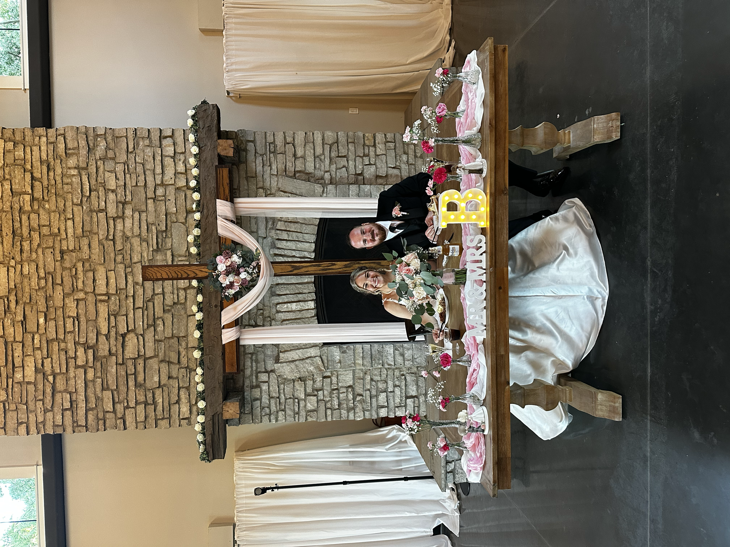 A man and woman at a wedding reception, seated at a decorated table with pink and white flowers, candles, and a large letter 'B' illuminated sign. The backdrop features a brick fireplace with a wood beam mantel decorated with flowers, and beige curta