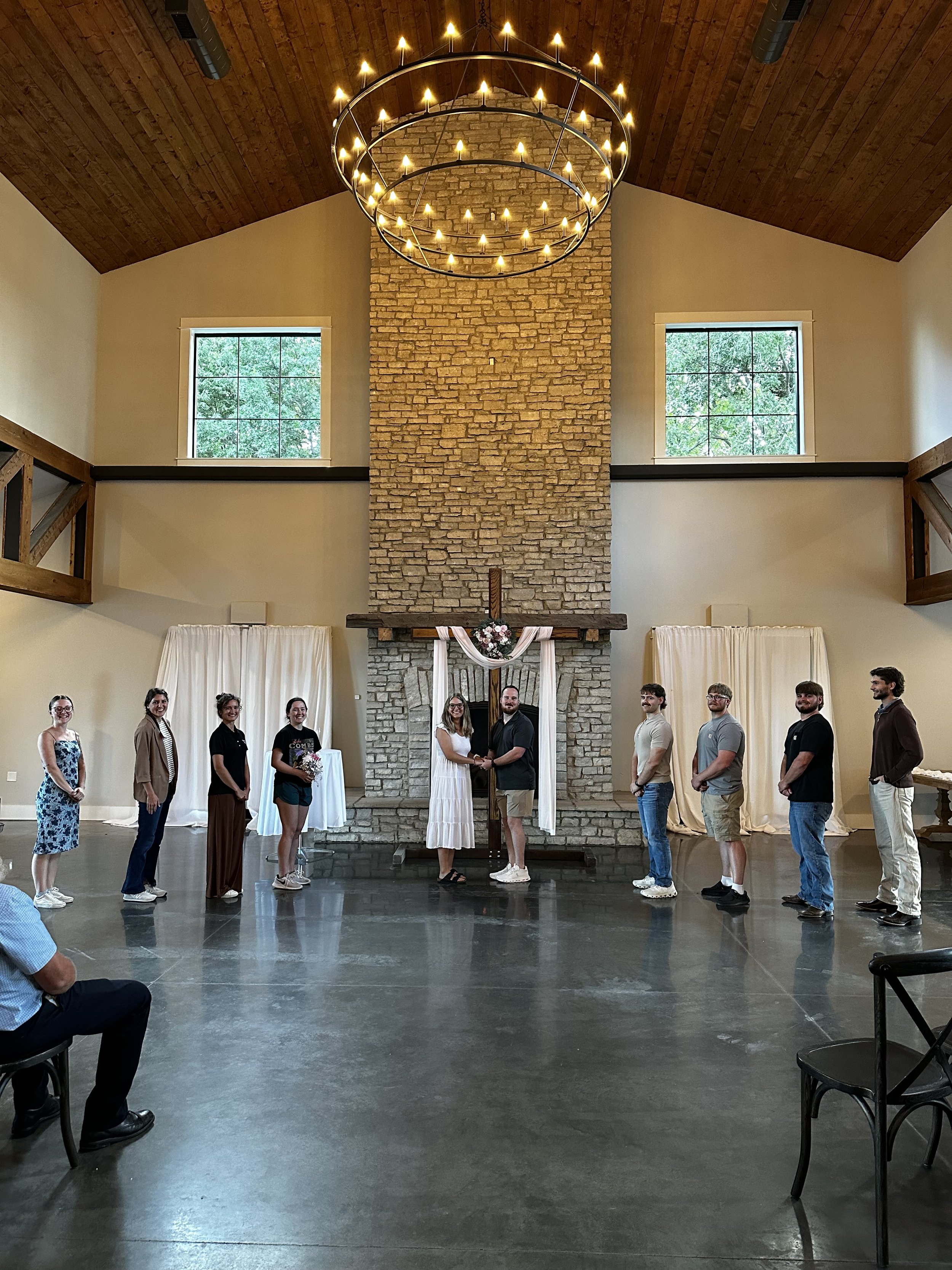 A group of people standing in a semi-circle inside a room with a stone fireplace, wooden ceiling, and chandeliers, celebrating a wedding with the bride and groom at the center holding hands.